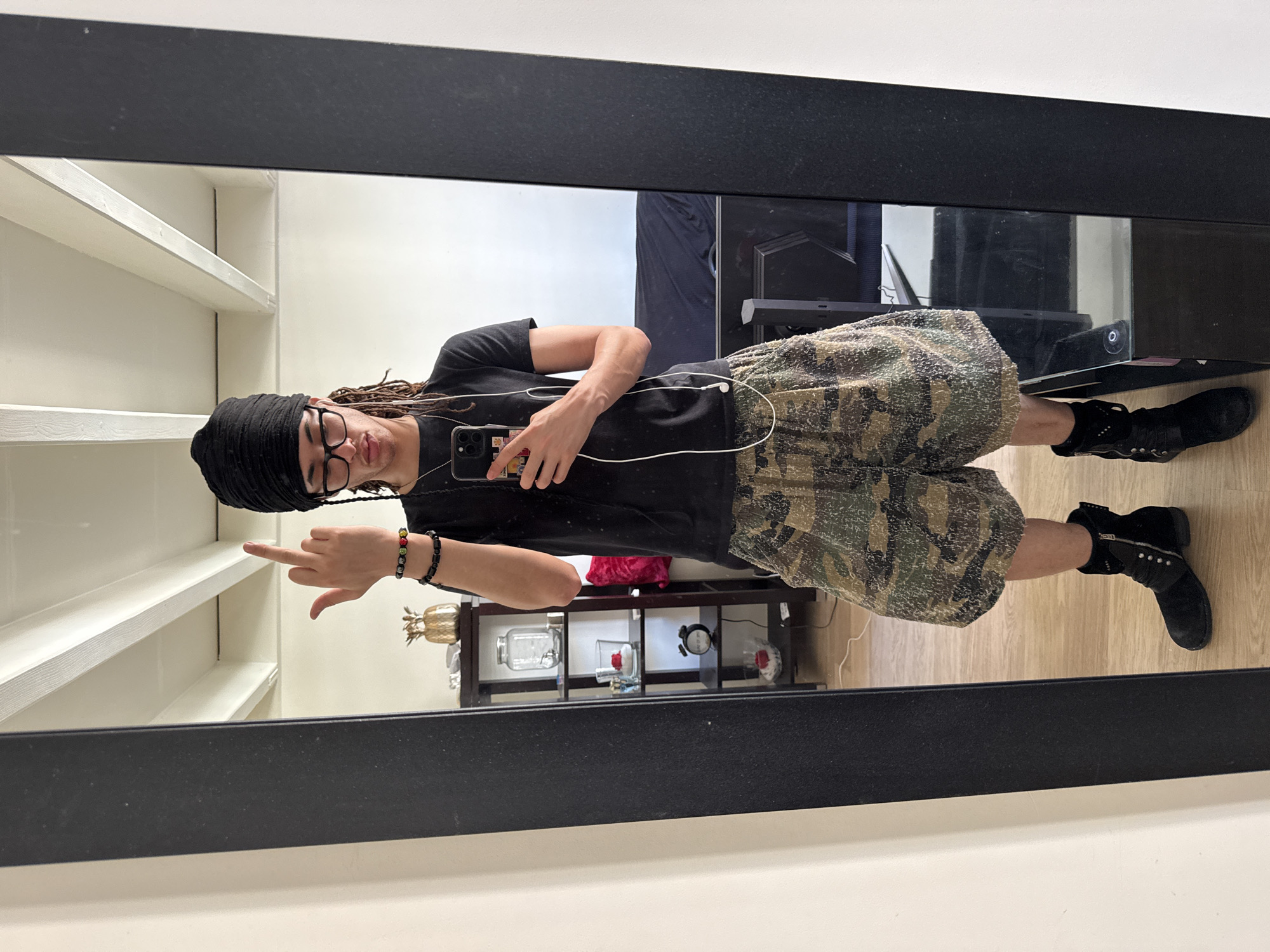 A person is seen in a full-body mirror selfie indoors. The person wears a black turban, glasses, a black t-shirt, camouflage shorts, and black boots. The person holds a phone in their right hand and is extending their left hand with the middle finger raised. Visible behind the person are white shelves, a black-painted door, and a black entertainment unit holding a TV and various decorative objects, including a pineapple figurine and a glass jar with a red rose inside. The floor appears to be wood. The location of the image is impossible to determine based on the information provided, therefore, Paris, France, cannot be confirmed.