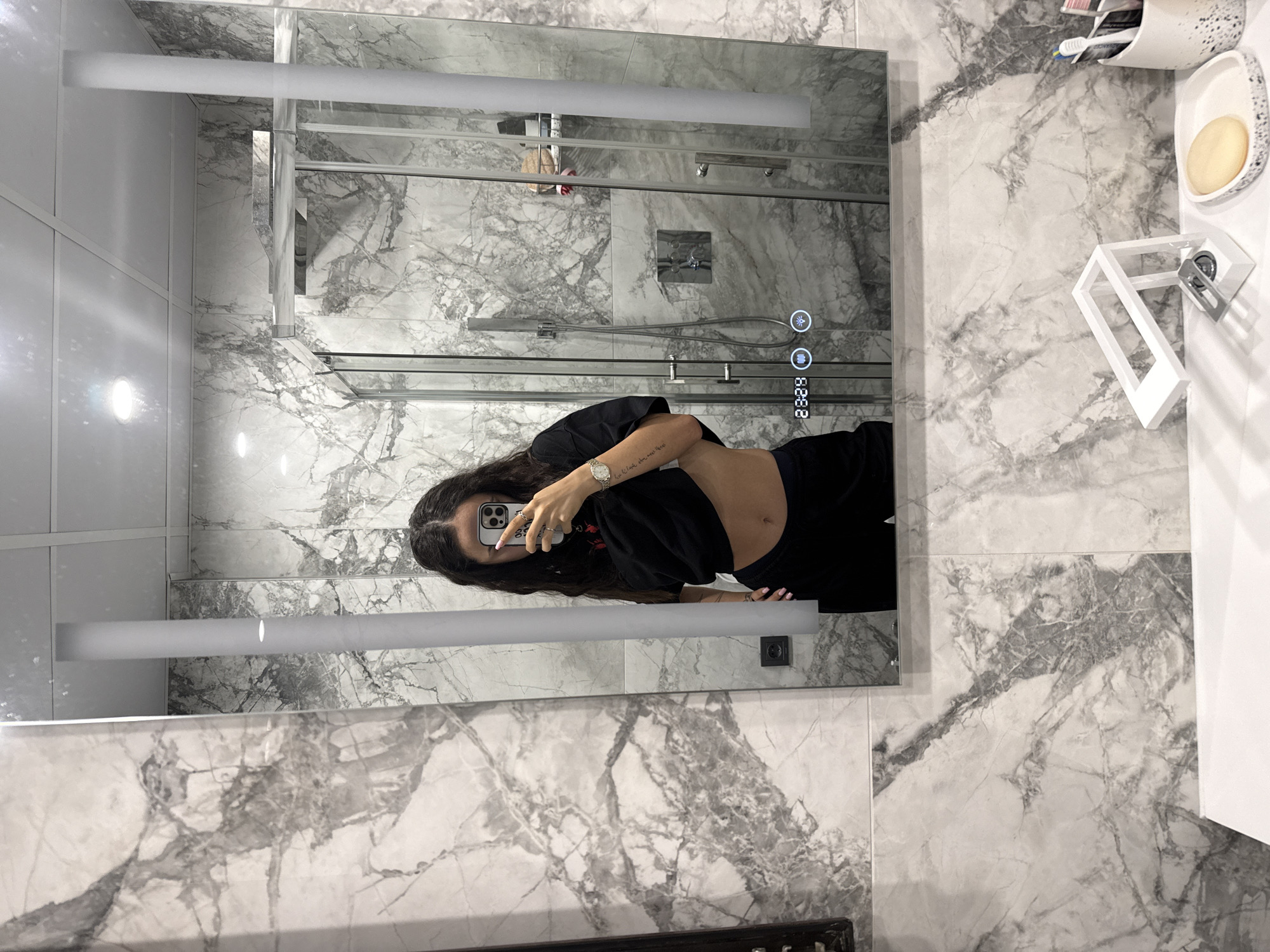 A woman takes a mirror selfie in a bathroom. She is standing in front of a vanity with a white sink and a modern white soap dispenser. Behind her, a large mirror reflects the bathroom, which features marble-patterned tiles on the walls and a glass-enclosed shower. The woman is wearing a black crop top and black pants. Her left arm is extended, holding a smartphone. A tattoo is visible on her left forearm. Her right hand is resting on her hip, and a watch is visible on her wrist. The mirror has a digital display showing the time "23:29" and icons for lighting and sound. The bathroom appears to be well-lit, with a showerhead and faucet visible in the reflection of the shower. The overall impression is that of a modern, stylish bathroom. There is no indication in the image that this location is in Shumen, Bulgaria.