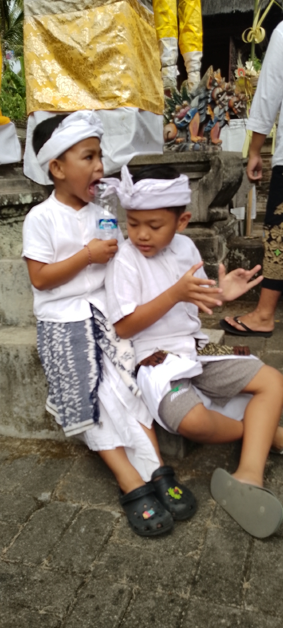 The image shows two young boys sitting on stone steps in what appears to be an outdoor cultural setting in Pekutatan, Indonesia. The boy on the left is wearing a white shirt, a patterned sarong, and black clogs. He is holding a plastic water bottle and has his mouth open as if speaking or reacting. The boy on the right is wearing a white shirt, grey shorts, and a patterned cloth around his waist. He is looking down and has his hands raised. Behind the boys, a yellow and white fabric is draped, and a stone carving of a creature is visible. The environment suggests a temple or a place of cultural significance.