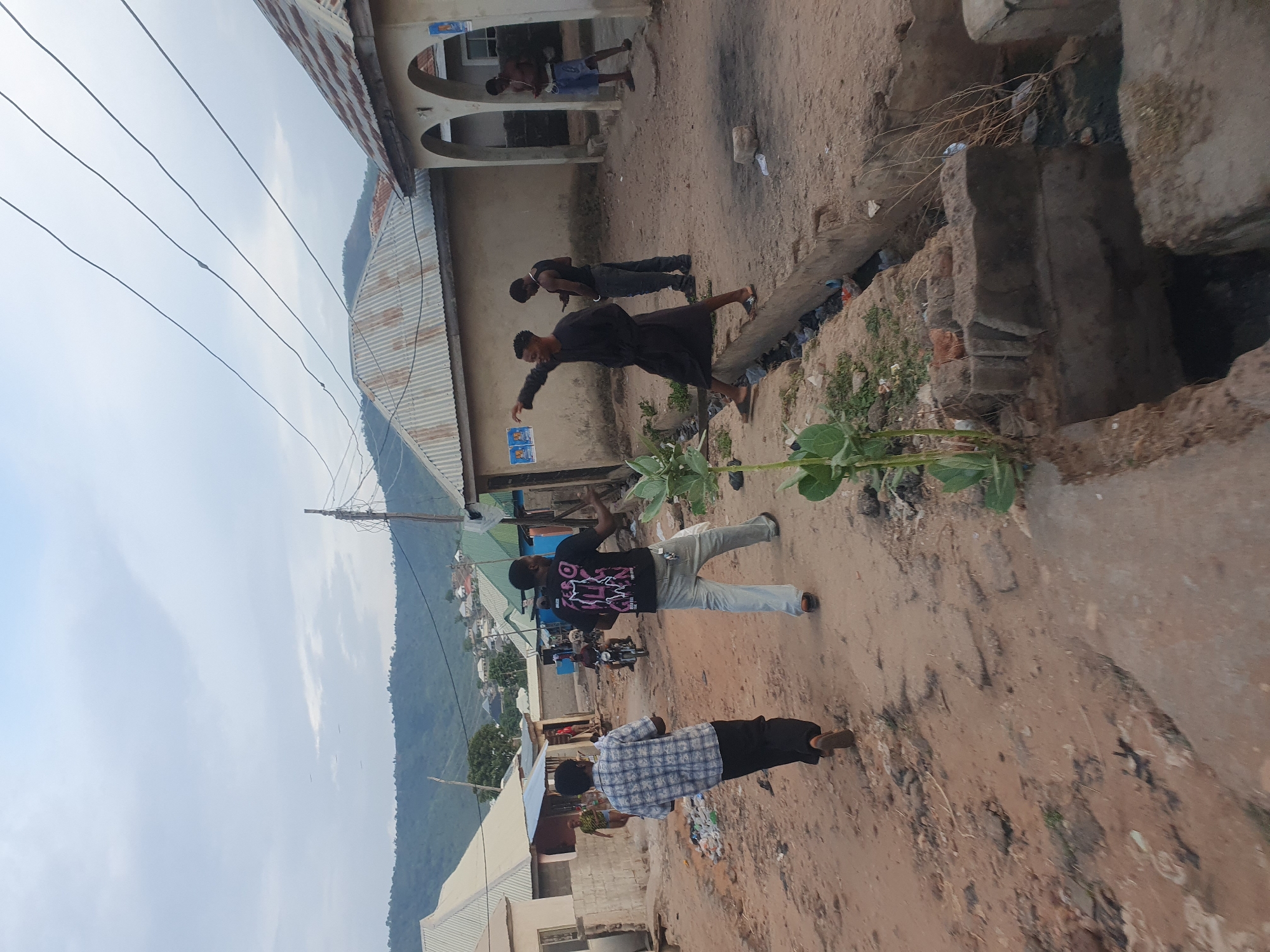 This media file captures an outdoor scene in what appears to be a residential or semi-urban area, likely in Lokoja, Nigeria, given the provided context. The image is oriented sideways, but depicts a dirt road or pathway with several people, buildings, and a mountainous background under a bright sky.
In the foreground, a man wearing a light blue and white plaid shirt and dark trousers walks towards the right. Further back, on the left, another man in a black t-shirt with a pink/purple graphic design (possibly reading "ZOOM" or "ZOOOM") and light-colored jeans stands, slightly raising his left arm. Adjacent to him, a man dressed in a long, dark robe or traditional garment gestures or points with his right arm, looking in the direction he is pointing.
To the right, a makeshift bridge or plank crosses over a ditch or drainage channel. Beyond this, near a light-colored building with arched entrances, two more men are visible. One is shirtless, wearing shorts, and the other wears a dark shirt and pants. They appear to be conversing or standing casually. In the far background, faint figures, possibly on a motorbike, are visible on a distant road, and lush green hills or mountains rise under a clear, light blue sky with some soft clouds.
The setting includes simple concrete buildings with corrugated metal roofs. Overhead power lines traverse the sky, and some green plants with large leaves grow near the ditch. The ground is unpaved, consisting of reddish-brown dirt, with some scattered debris. The bright, clear daylight suggests it is either mid-morning or late afternoon. No strong emotions are discernible on the faces of the individuals.