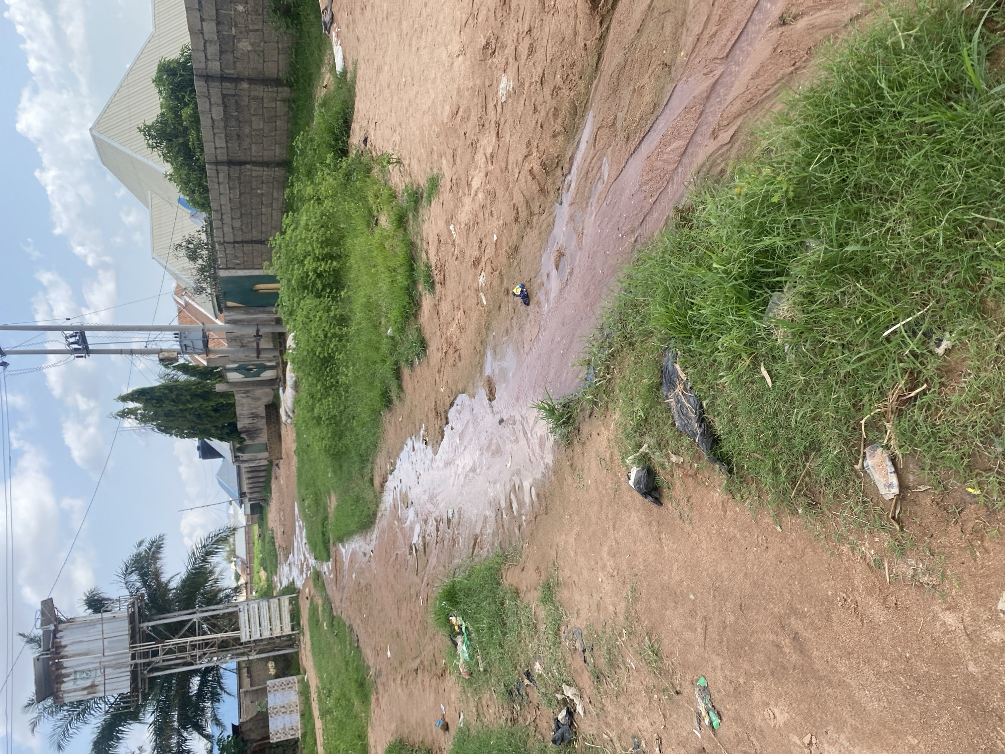 This image depicts an outdoor scene in Jauro Yinu, Nigeria, characterized by a muddy, water-filled trench running through a predominantly sandy area. The trench is flanked by patches of overgrown green grass. The ground is littered with various debris, including plastic bags and bottles, suggesting a lack of proper waste management.
In the background, there is evidence of human settlement. A stone wall separates the sandy area from a street or pathway, beyond which are buildings with light-colored, sloped roofs and some trees. A prominent structure in the mid-ground is a large, metal water tank mounted on a metal framework, a common sight in areas with unreliable water supply. Electrical poles and wires are also visible, indicating the presence of infrastructure.
The weather appears to be bright and sunny, with a clear blue sky dotted with white clouds. The lighting suggests it is daytime. There are no people visible in the frame, and the overall scene conveys a sense of a developing or possibly impoverished area, given the visible waste and infrastructure elements like the water tank. The mood is neutral, focusing on the physical environment and its condition. No discernible text is present in the image.