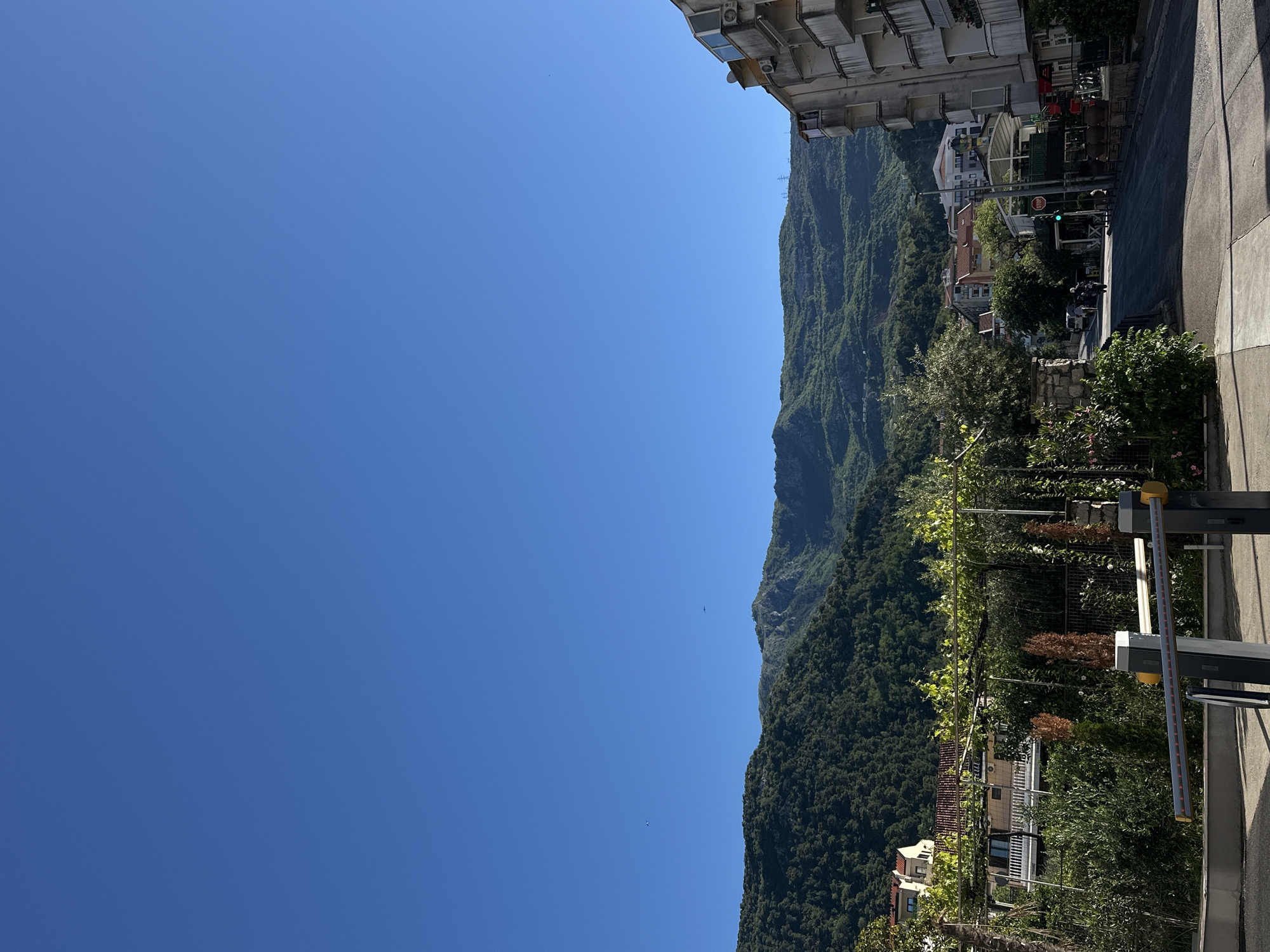The image shows a sunny, clear day in Budva, Montenegro, with a focus on the landscape. A large, verdant mountain dominates the right side of the frame, its slopes covered in dense forest. Closer to the viewer, there are houses and buildings nestled among the trees. A sidewalk and a security barrier are visible in the foreground. A glimpse of urban development, including buildings and a road, is seen further along the right edge. The left side of the image is a clear, bright blue sky. A bird is flying in the sky.