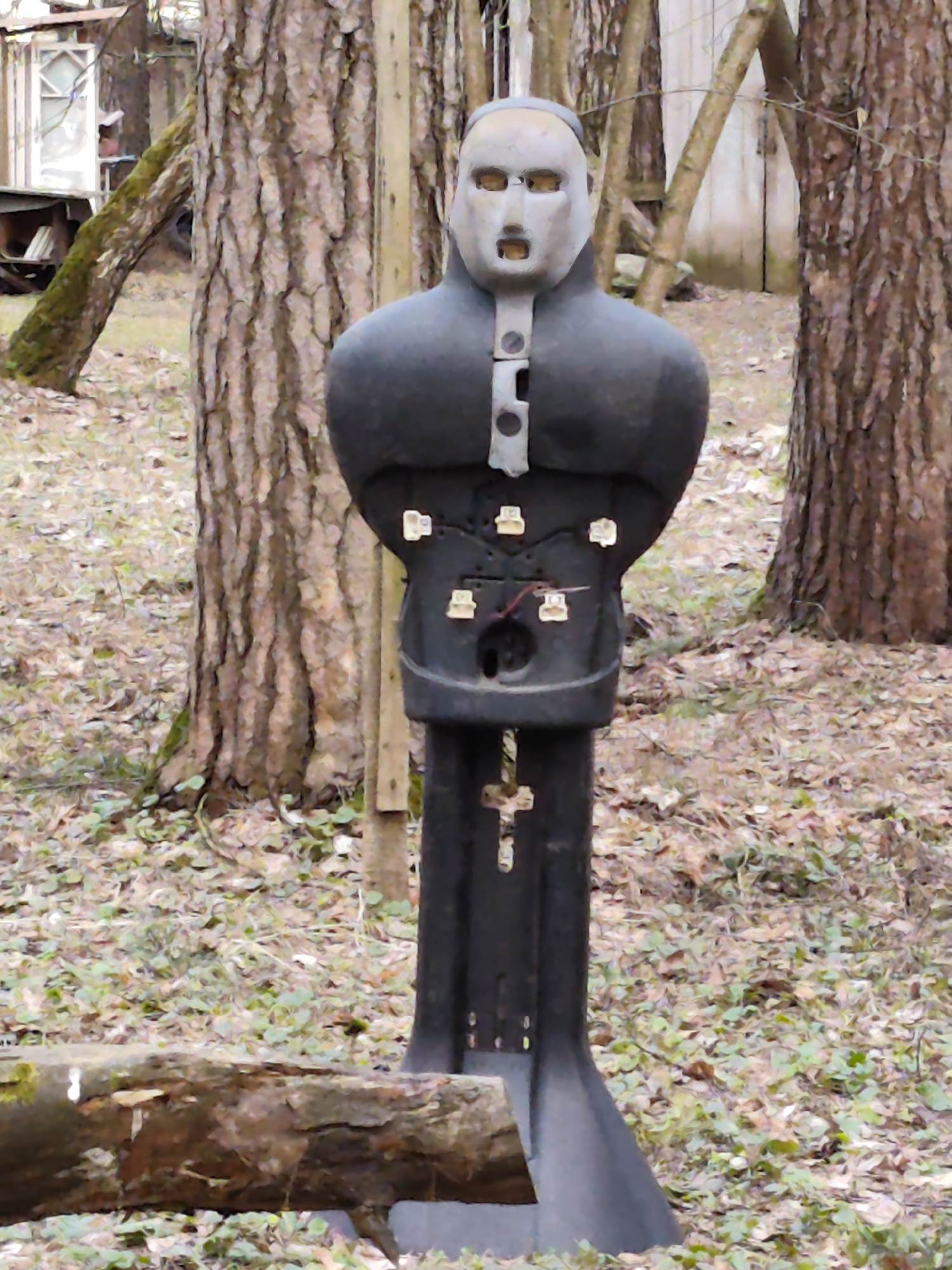 The image shows a weathered, human-shaped target mannequin standing outdoors in a wooded area, likely Kratovo, Russia, given the request. The mannequin has a grayish-white head with cut-out eye and mouth holes, and a black, padded torso. Several small, white plastic objects are attached to the torso, and wires protrude from a hole in the abdomen. It's standing among fallen leaves and several tree trunks. A partially visible log rests at the bottom of the frame. In the background, a building or structure is visible.