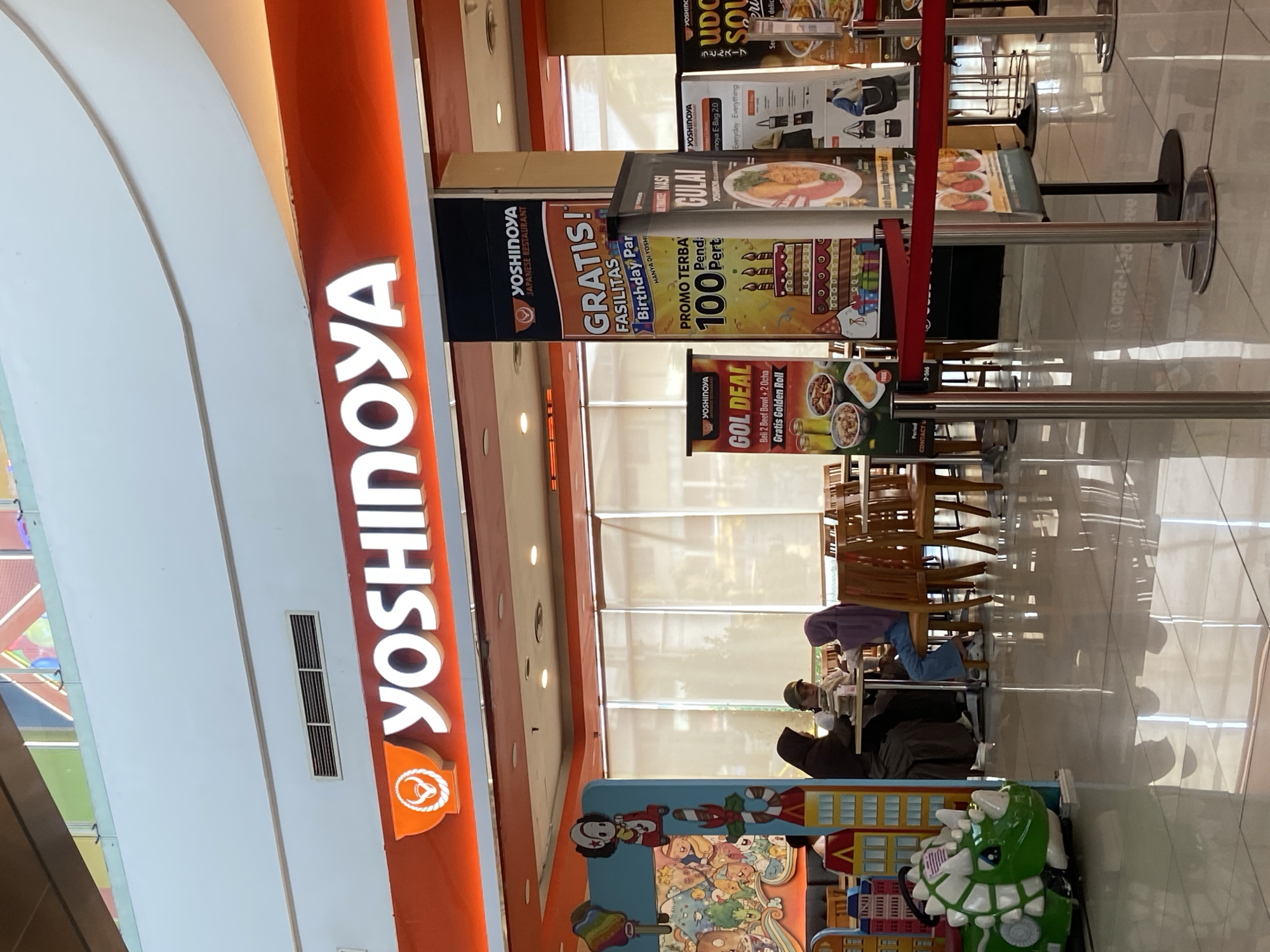 The image captures the interior of a Yoshinoya restaurant, likely located in Kudus, Indonesia, given the prevalence of Yoshinoya franchises in that region. A large, illuminated Yoshinoya logo with an orange background is prominently displayed above the entrance. To the left, a colorful children's play area features a green dinosaur ride.
Inside, three individuals are seated at a table, appearing to be a family, with two adults and one child. They are engaged in conversation or dining. The restaurant interior features wooden tables and chairs, and the floor is tiled and reflective, suggesting a well-maintained space.
Several promotional banners are displayed in the foreground, advertising various Yoshinoya deals and offers, including "GOL DEAL" and birthday party facilities. The scene is brightly lit, likely by overhead lighting within the mall or building where the restaurant is situated. The overall impression is that of a casual dining environment within a commercial space.