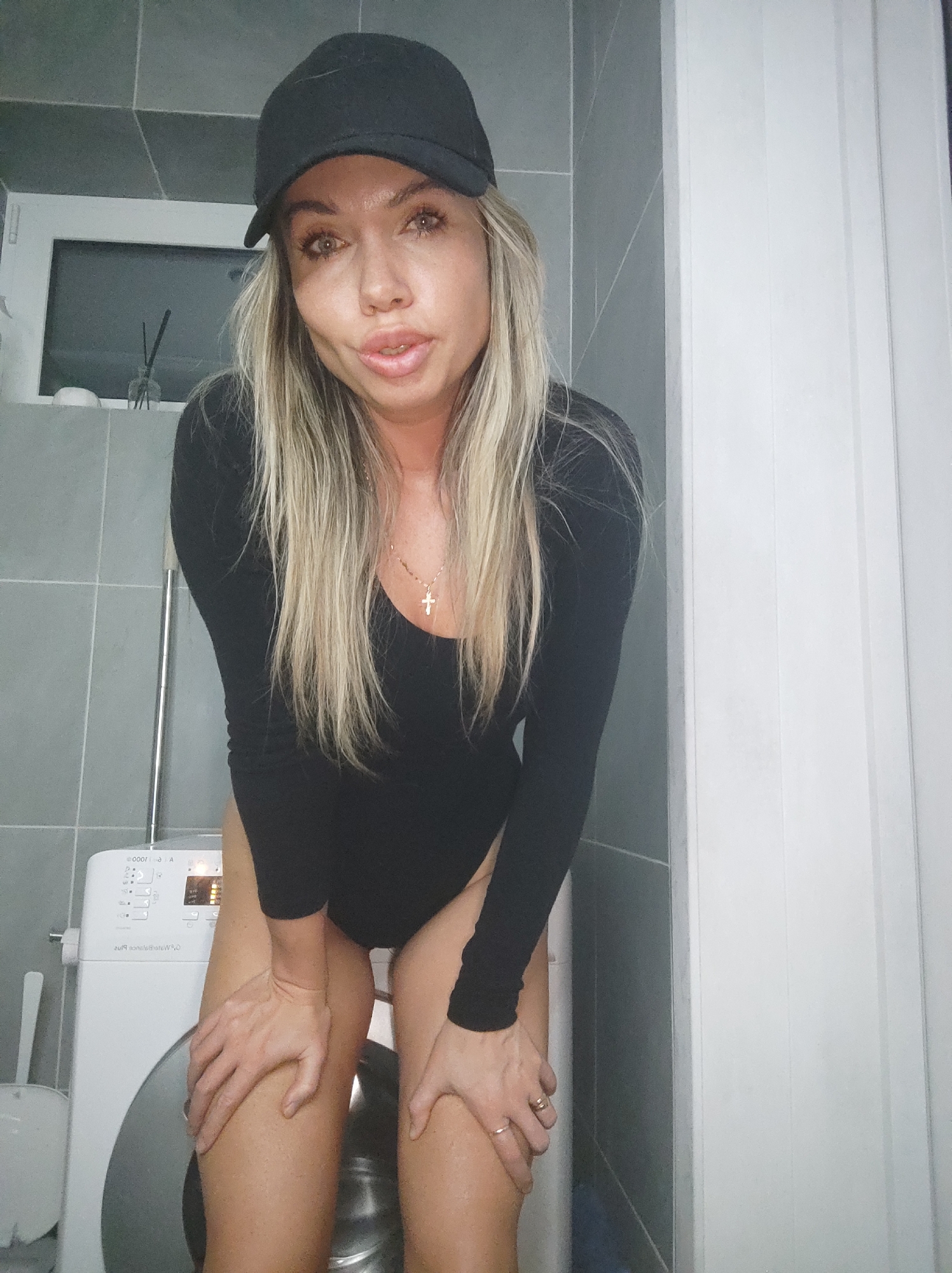 A woman with blonde hair and a black cap is posing in a bathroom. She is wearing a black bodysuit. Her hands are on her knees, and she is looking at the camera. In the background, there is a washing machine and a window with reeds in a vase. The bathroom has gray and white tiles. The location is not specified to be Votkinsk, Russia.
