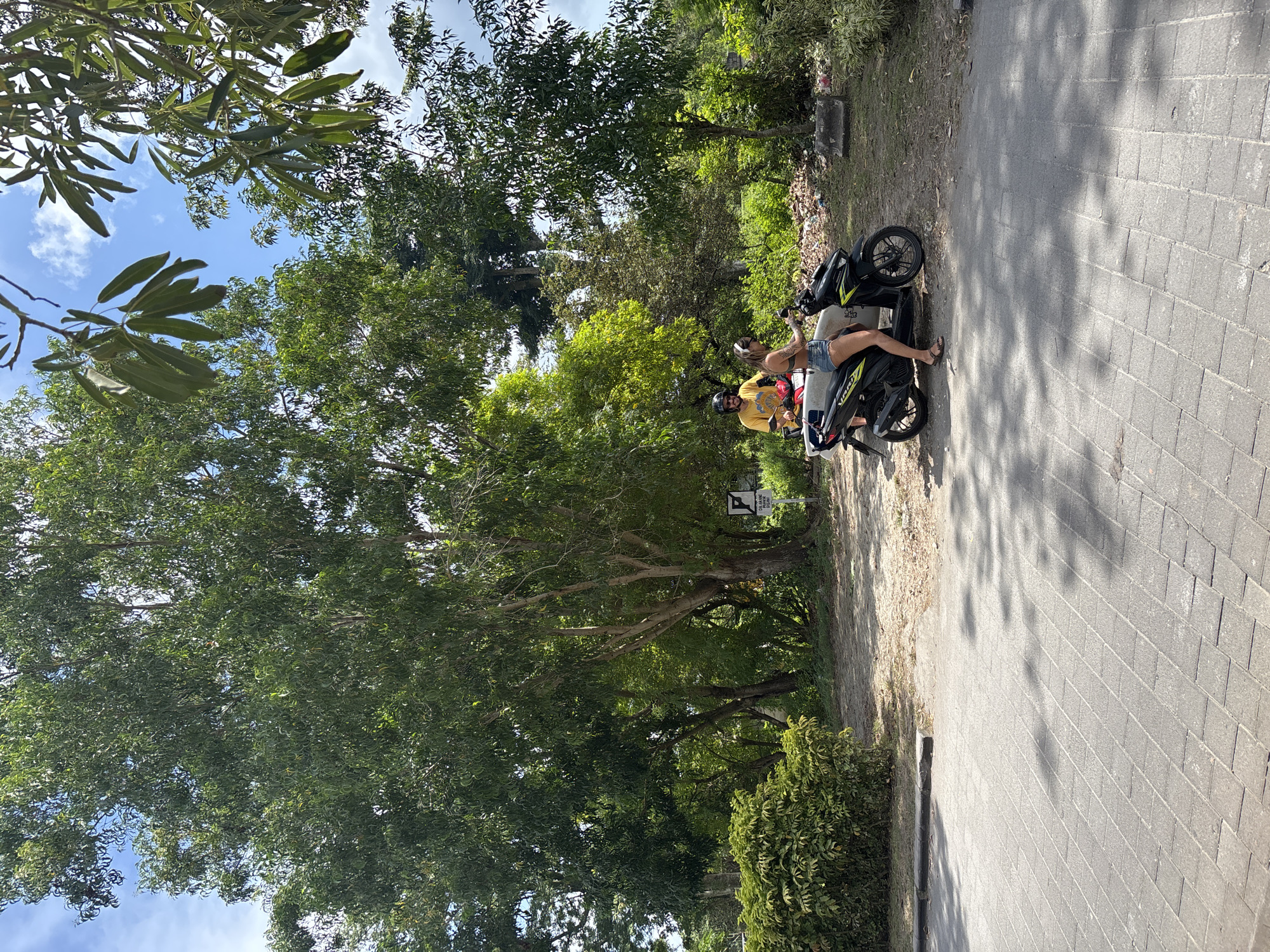 Two individuals are present, sitting on a black scooter parked along a paved walkway adjacent to a tree-lined area. The location is Kuta Selatan, Indonesia. One individual is wearing a yellow shirt and helmet, the other appears to wear a tank top and shorts. Vegetation includes trees of varying sizes and a small bush along the walkway's edge. Pavement stretches from the scooter to the right edge of the image.