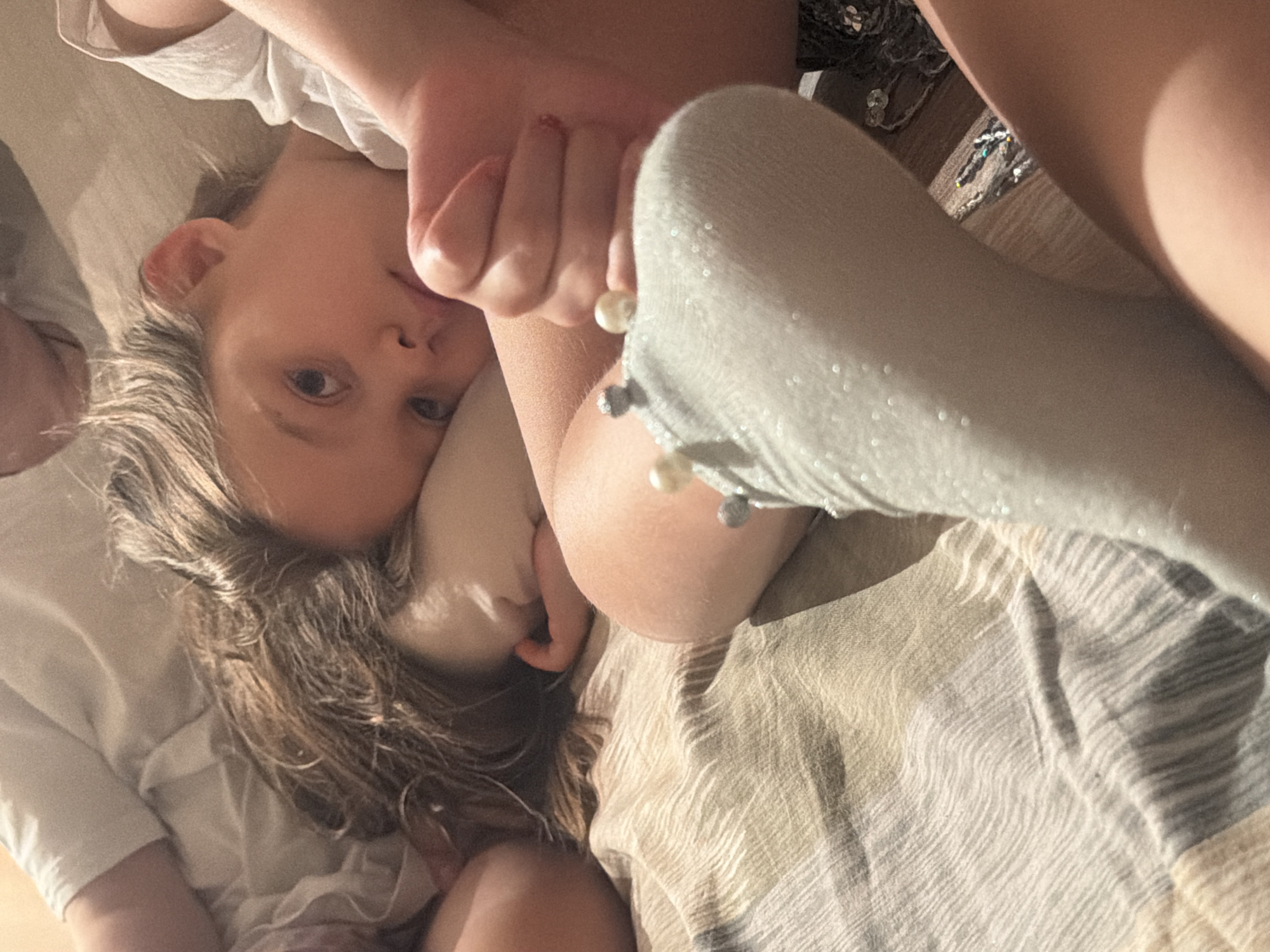 The image depicts two children lying on a striped bedspread. The child closer to the camera has blondish hair, blue eyes, and is partially obscured by a foot wearing a sparkly, light gray sock adorned with pearls. The other child, lying below, has brown hair. They are lying in an undefined place, and there is no indication of the location being Krasnoyarsk, Russia.