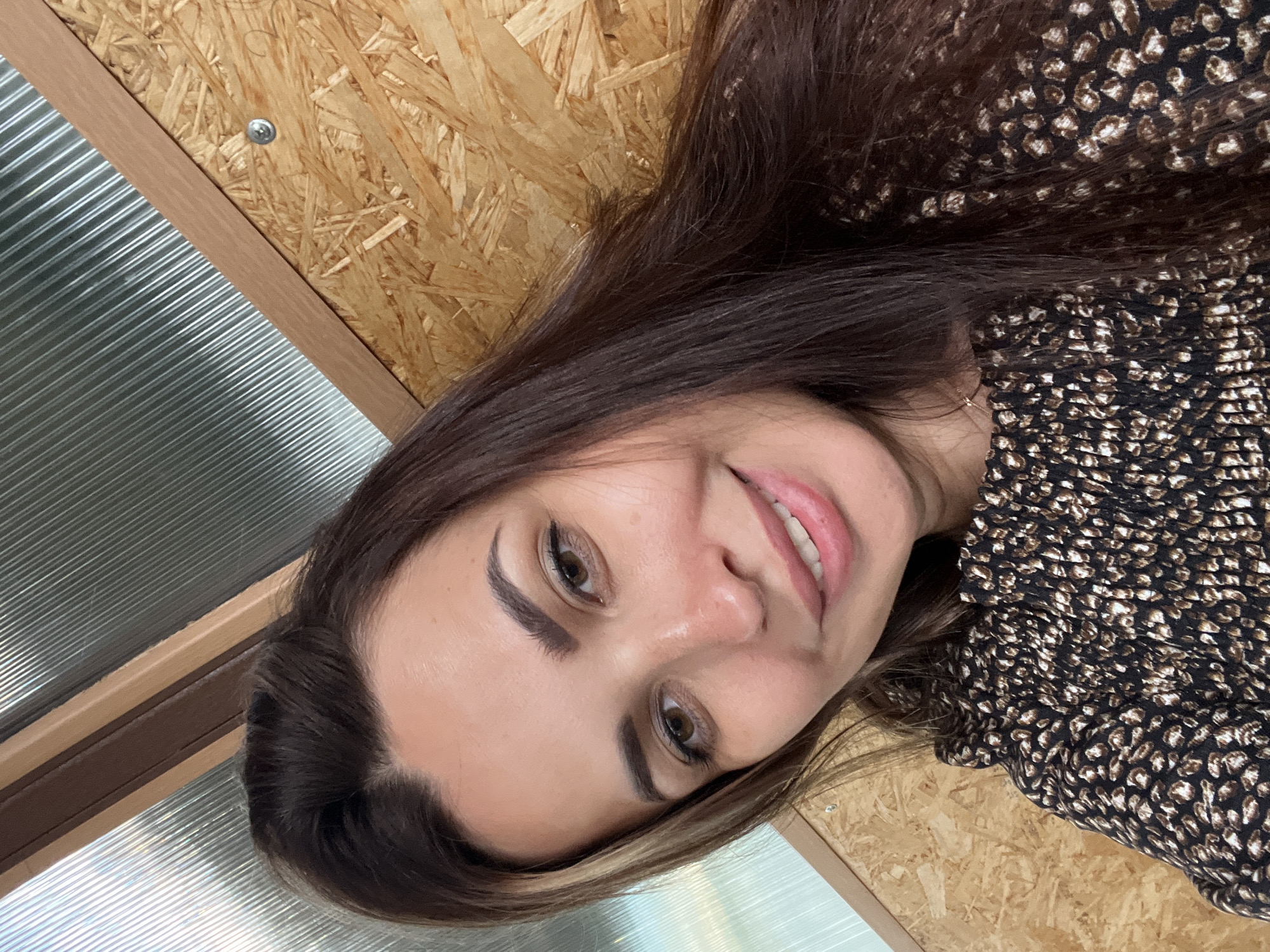 An outdoor, close-up, selfie-style photograph shows a woman with long, dark brown hair and brown eyes. She is smiling at the camera. She is wearing a dark-colored top with a leopard-print pattern. The background consists of a wooden wall and a corrugated metal ceiling. The lighting is natural.