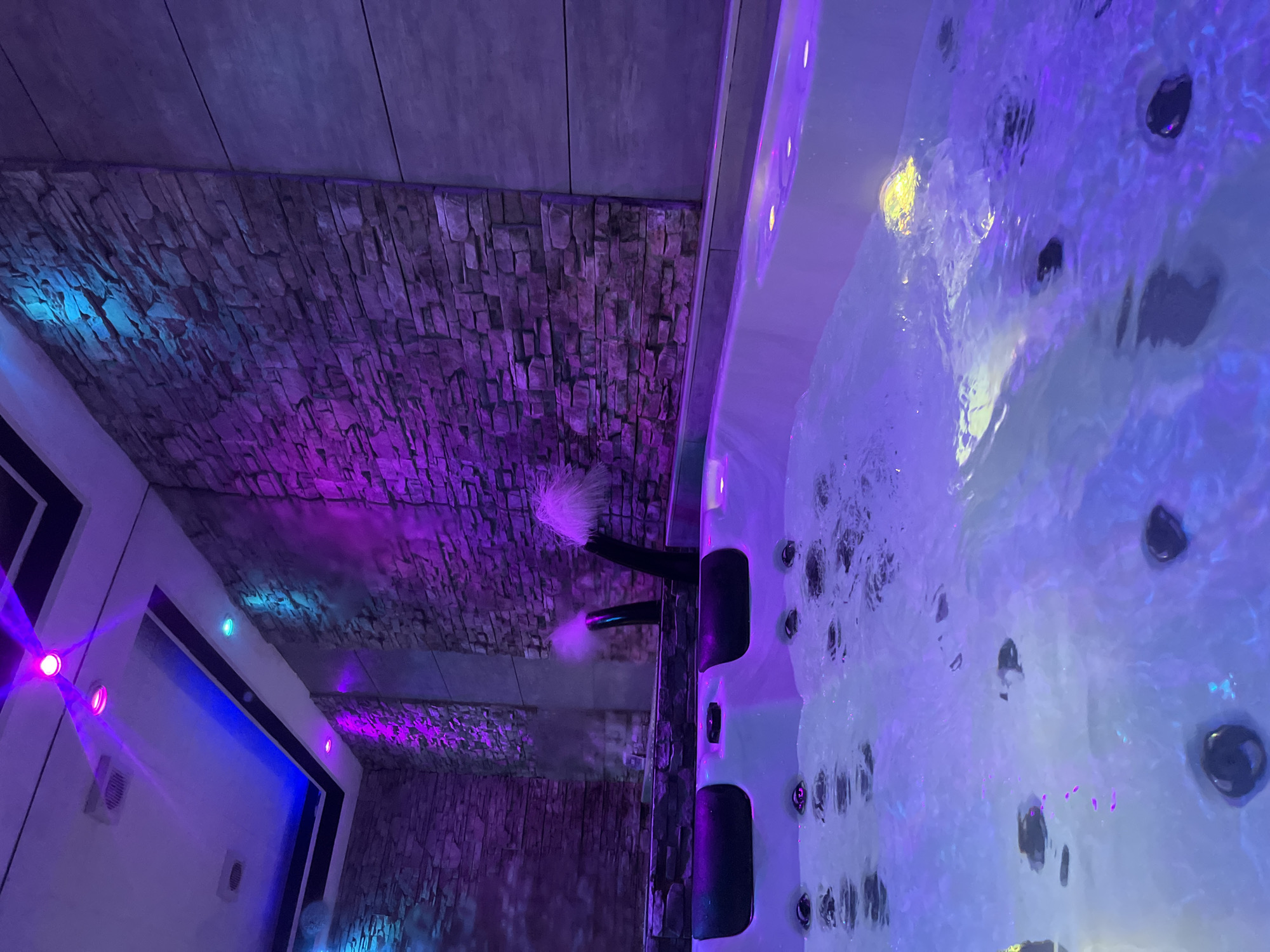 The image shows an indoor hot tub illuminated with purple and blue lights. Two black spouts extend from a stone wall into the tub, emitting streams of water that create a feathery effect at the ends. The water in the hot tub is agitated and appears bubbly. The surrounding walls are textured with stone and wood paneling. On the left side of the image, a doorway with colored lighting is visible. The location is in City of Zagreb, Croatia.