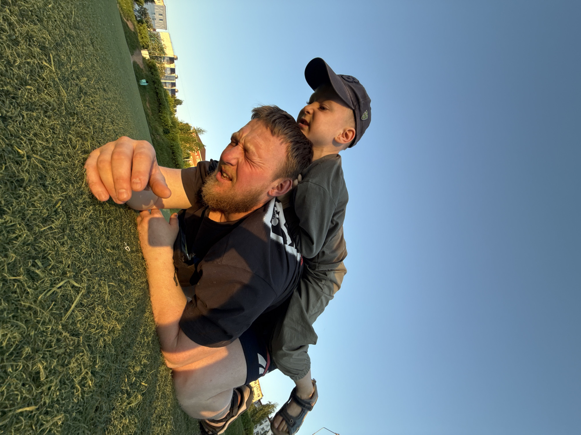 A father lies face-down on artificial turf while his young son sits on his shoulders. The father is fair-skinned with a beard and is wearing a black t-shirt and shorts. The son has fair skin and is wearing a baseball cap, a long-sleeved shirt, and sandals. The picture seems to be taken from ground level, as the father is looking to be grimacing from the weight on his back. The location is likely a sports ground with buildings visible in the background, possibly in Chaikovsky, Russia. The sky is light blue.