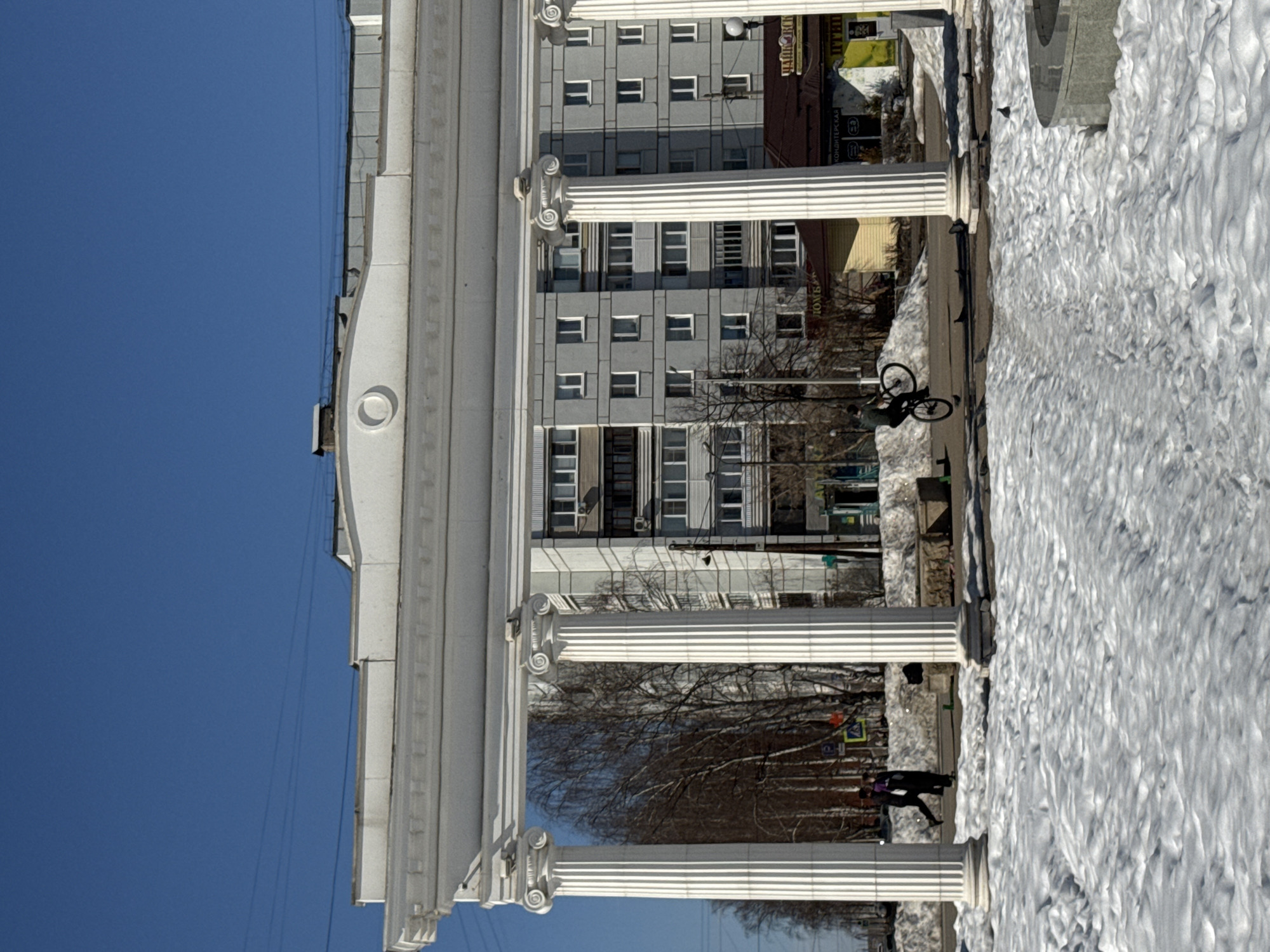 The image depicts an outdoor urban scene in Chaikovsky, Russia, under a clear blue sky, characterized by melting snow. In the foreground, the ground is covered with lumpy, textured snow. A gray, circular stone fountain basin is partially visible on the bottom right, also surrounded by snow.
Dominating the mid-ground is a white, classical-style archway featuring four visible fluted columns with Ionic capitals, supporting a plain entablature. On the paved surface beneath the archway, a male individual in dark clothing is performing a wheelie on a black bicycle, balanced on its rear wheel. To the left of the bicyclist, two other individuals walk away from the viewer; one wears a dark coat with a purple item on their back, and the other wears a dark coat. Several pigeons are scattered on the paved ground near the columns and bicyclist.
Behind the archway, patches of snow accumulate along what appears to be a street or pathway. Bare deciduous trees are visible on the left and center-left. In the background, a multi-story, light-gray residential building with numerous rectangular windows and dark balconies is prominent. To the right of this building, lower-rise commercial structures with brown roofs are visible. One sign clearly displays "КОНДИТЕРСКАЯ" (Confectionery), and another partially visible sign includes "ЧАЙКОВСКИЙ", confirming the city. Utility poles and overhead power lines are visible throughout the background.