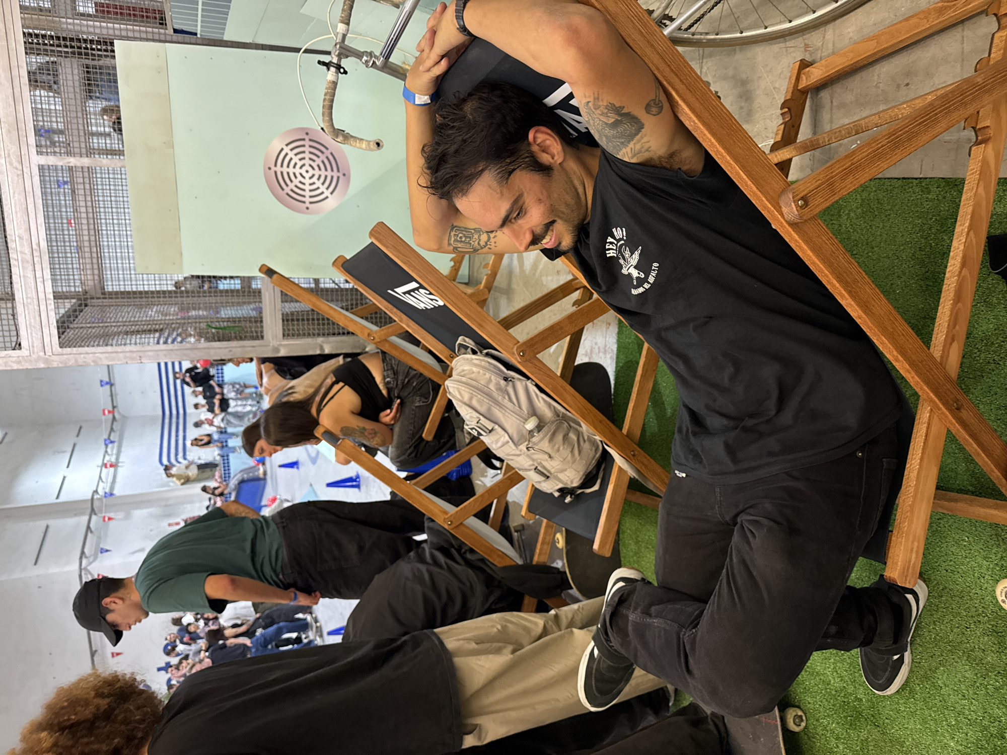 The image depicts a group of people relaxing on beach chairs in what appears to be an indoor recreational area, likely located in L'Hospitalet de Llobregat, Spain. A man with dark hair and a mustache, wearing a black t-shirt and black jeans, is reclining in a chair in the foreground, with his arms behind his head. He has visible tattoos on his arms.
Behind him are other individuals sitting in similar chairs, some with backpacks. A skateboard is visible under one of the chairs. The background includes a light green wall with a circular ventilation grille, and a glimpse of a bicycle. The flooring is partially covered with artificial turf. In the far background, more people are visible, and some orange and blue traffic cones are located on the floor next to a swimming pool. A barred ceiling structure is also visible.