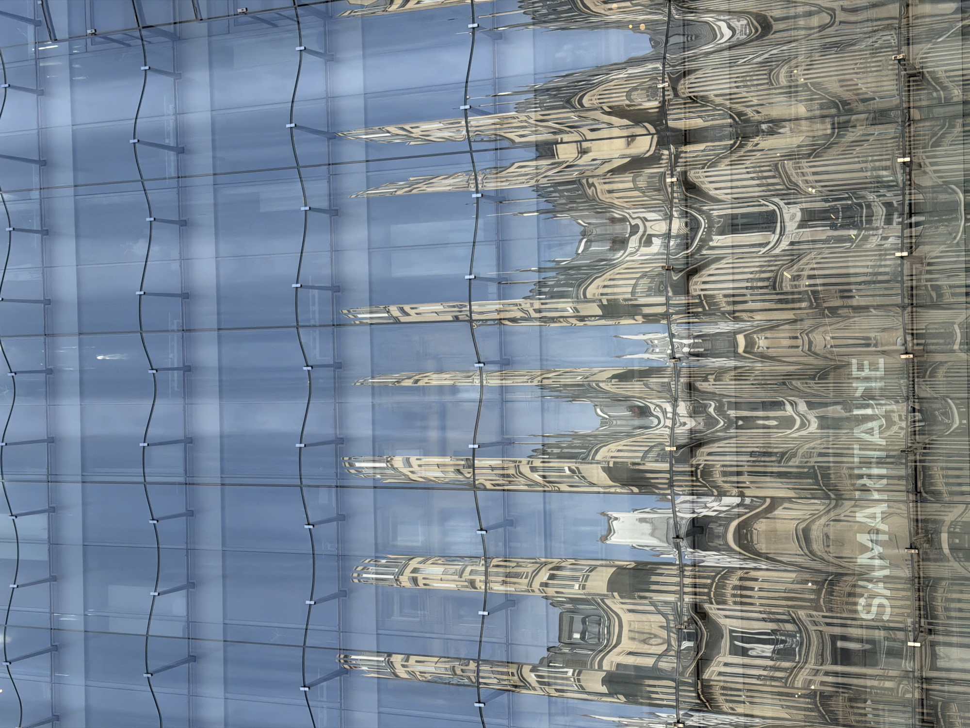 This image captures a close-up of a modern, multi-paneled glass facade, showcasing its intricate structure and reflective properties in Paris, France. The facade is composed of numerous rectangular glass panels, arranged in a grid and supported by horizontal metallic beams. Running vertically across the facade are thin, undulating tension cables or rods, secured by small, square silver fixtures attached to the glass panels.
Reflected prominently in the wavy surface of the glass is a grand, classical-style building, characterized by its ornate architecture, numerous windows, and decorative cornices. The reflection is significantly distorted, stretching and warping the classical building's features into abstract, undulating patterns. Patches of light blue sky are visible in the reflection between the architectural elements. The interplay between the sleek, modern glass and the distorted, traditional reflection creates a visually dynamic effect.
In the lower right quadrant of the reflected building, the word "SAMARITAINE" is clearly visible in white capital letters. The scene is brightly lit by daylight, indicating a clear weather day. The image highlights the juxtaposition of contemporary and historical architecture typical of a city like Paris.