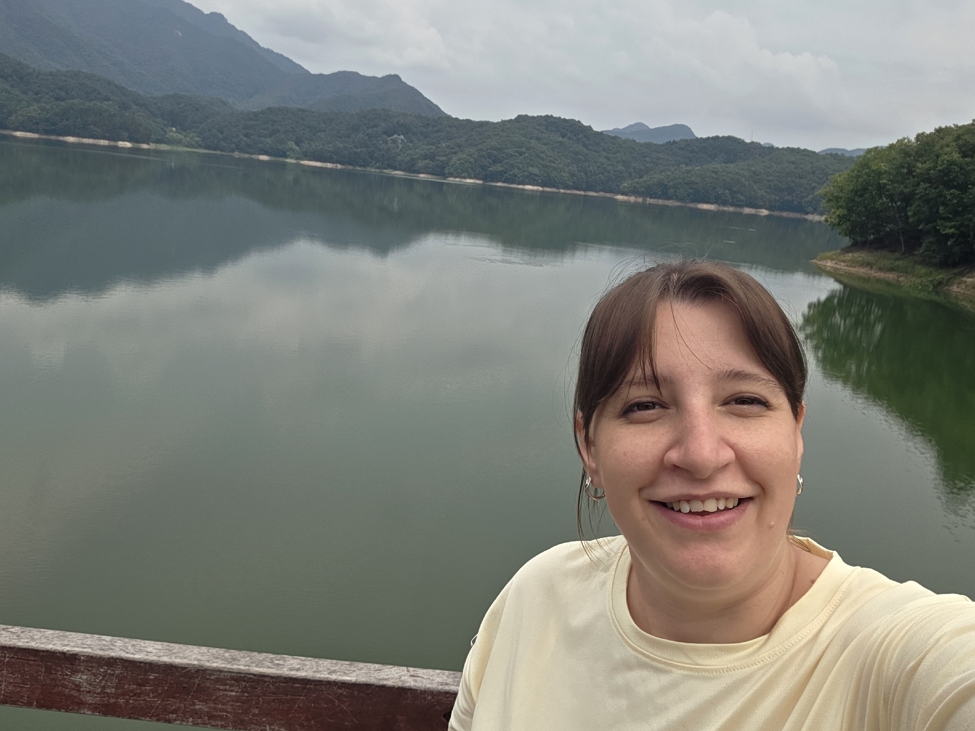 A woman smiles into the camera, taking a selfie. She is wearing a light yellow t-shirt and silver hoop earrings. Behind her, a large, calm body of water reflects the overcast sky and the forested mountains on the opposite shore. A weathered wooden railing runs along the bottom of the frame, in front of the woman and the water. The scene appears to be at a reservoir or lake in Gapcheon-myeon, South Korea, given the natural landscape.