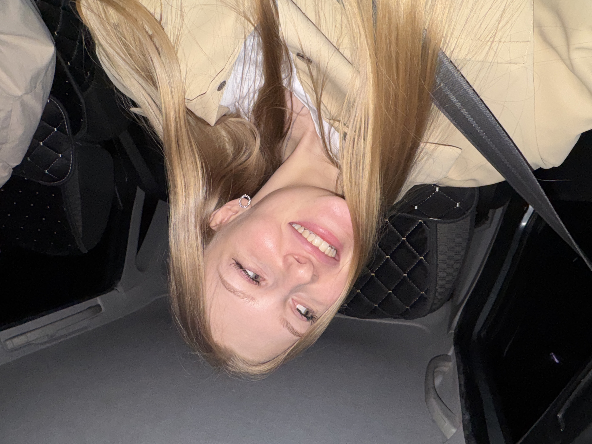 A fair-skinned woman with light blonde hair is inside a car, wearing a tan jacket over a white shirt and a seatbelt. She is smiling and facing the camera. The car's interior features black quilted seats. The location is likely Perm, Russia, based on context, but there is no specific detail confirming this.