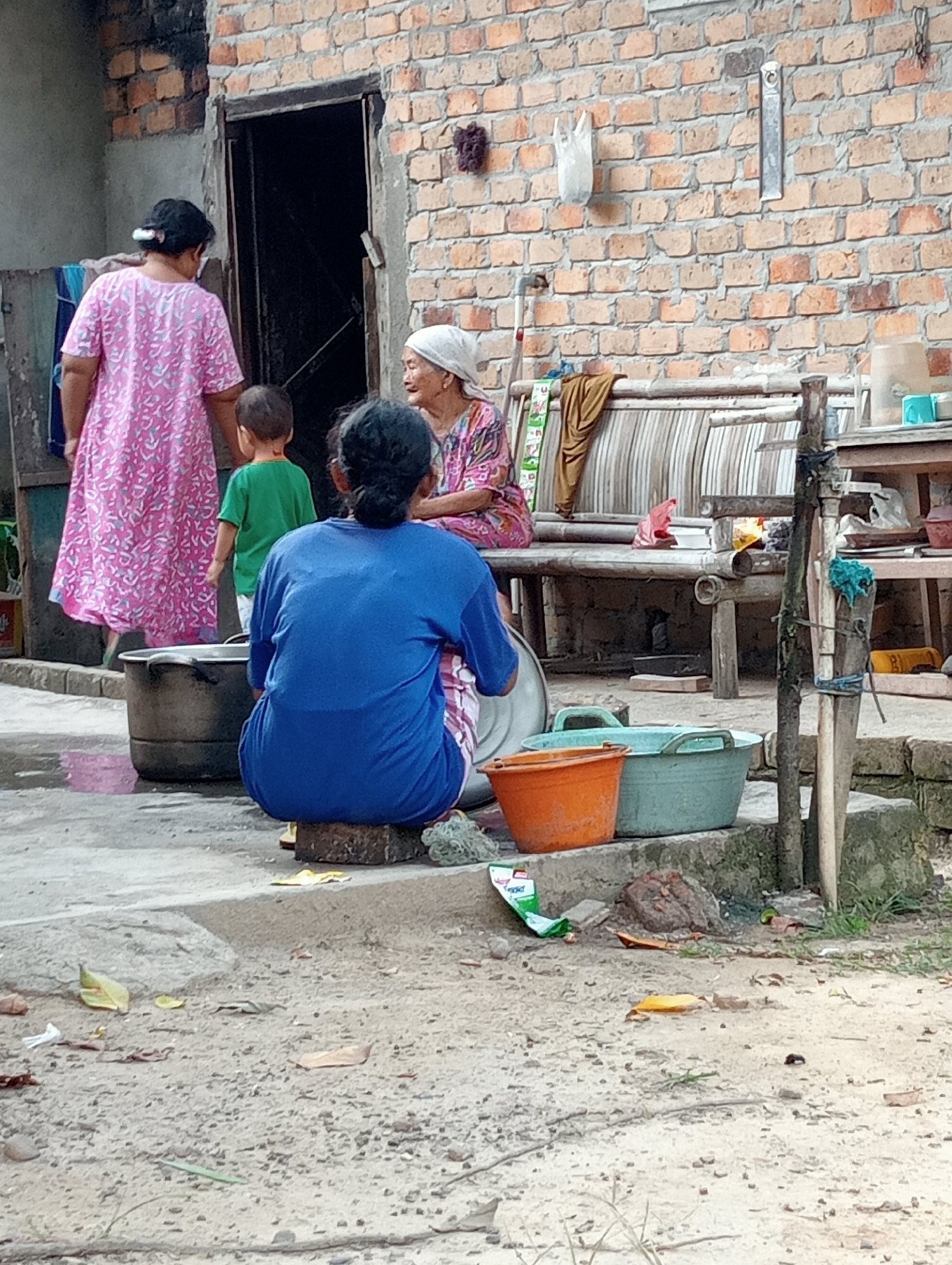 The image captures an outdoor scene in Kota Negara Timur, Indonesia, focusing on a group of individuals gathered near a brick building. In the foreground, a person is seated on a brick, facing away from the camera, wearing a blue long-sleeved shirt. To the left, a large metal pot is visible. Further back, a woman in a pink dress is standing, holding the hand of a young child in a green shirt. An elderly woman wearing a headscarf and a patterned shirt is seated on a wooden bench in the background, interacting with another person. A rustic wooden bench is situated behind the seated elderly woman, with various items on and around it. The environment appears to be a courtyard or alleyway, with a rough, textured ground surface. The brick building in the background has an open doorway. The overall impression is a candid snapshot of daily life in a residential area.