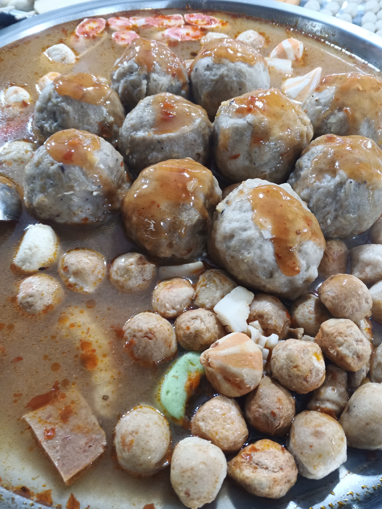 A close-up, overhead shot captures a large, circular metal basin filled with various types of meatballs and other ingredients submerged in a flavorful, reddish-orange broth. The meatballs are diverse in size and texture. Some are large, smooth, and coated with a glossy sauce that appears to be chili-based, while others are smaller and have a more textured, irregular surface. Mixed in with the meatballs are slices of pale, rectangular food item, possibly tofu or a similar product, and some lighter-colored, possibly white, pieces of food. There are also some pink and white striped objects, likely fish cakes or processed seafood. The broth itself has small flecks of red and orange, indicating the presence of spices or chili. The overall impression is that of a prepared dish, likely a soup or stew, ready for serving. The setting appears to be a food stall or restaurant, possibly in the city of Sukabumi, Indonesia, given the commonality of such dishes in Indonesian cuisine. There are no people or distinct environmental features visible beyond the contents of the basin and the basin itself, which has a slightly reflective, metallic rim.