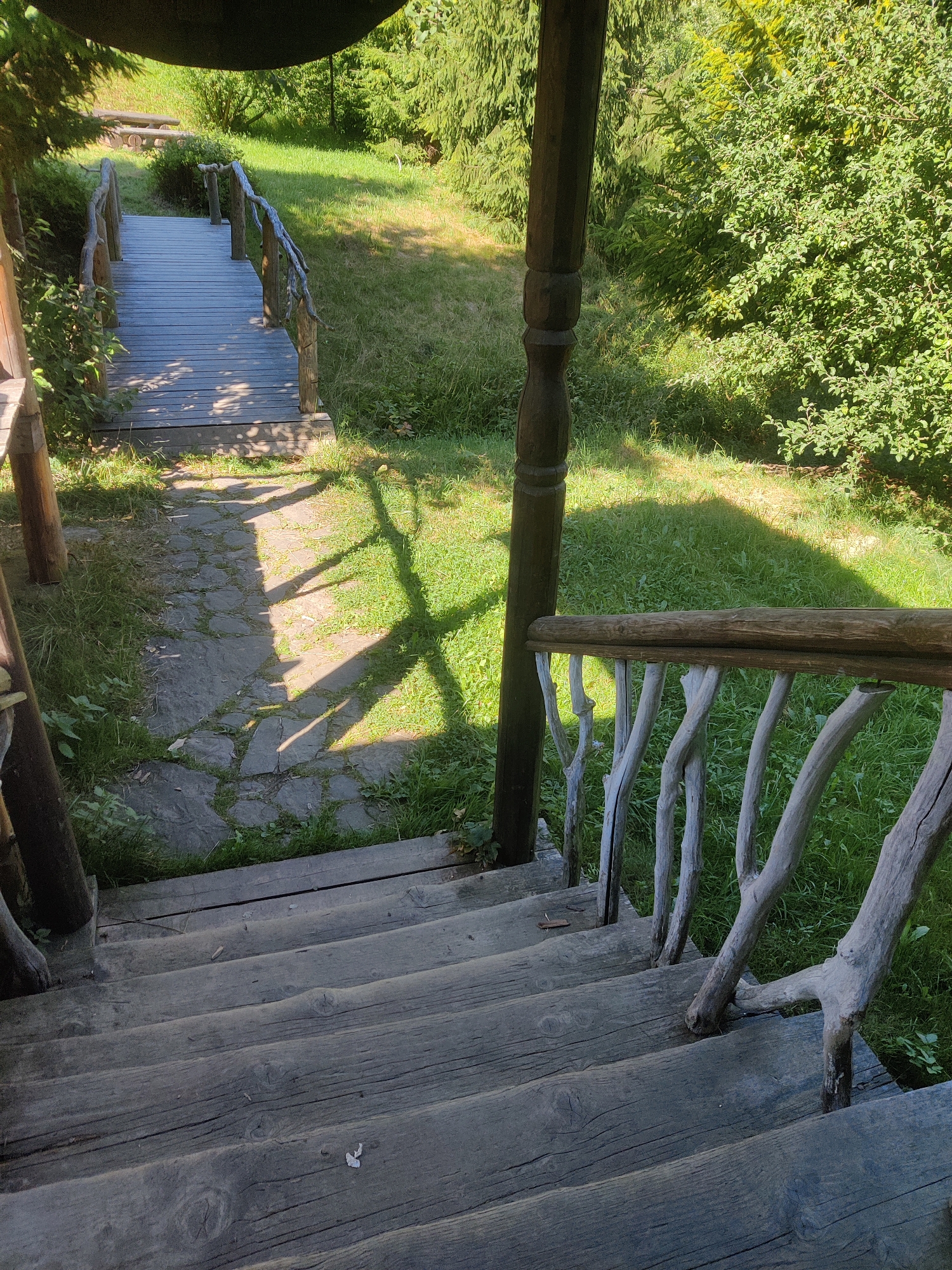 This image depicts a rustic wooden staircase leading downwards from a covered porch area. The staircase is made of rough-hewn wooden planks, showing signs of age and weathering. The railing is constructed from irregularly shaped branches, giving it a natural, organic look. To the left of the stairs, a weathered wooden plank pathway leads towards a wooden bridge. Beyond the bridge, a grassy area is visible, with scattered trees and lush green foliage in the background, suggesting a natural or park-like setting. The scene is illuminated by dappled sunlight, casting shadows on the ground and the wooden structures, indicative of an outdoor environment on a sunny day. The location is identified as Izky, Ukraine. There are no people or animals present in the image.