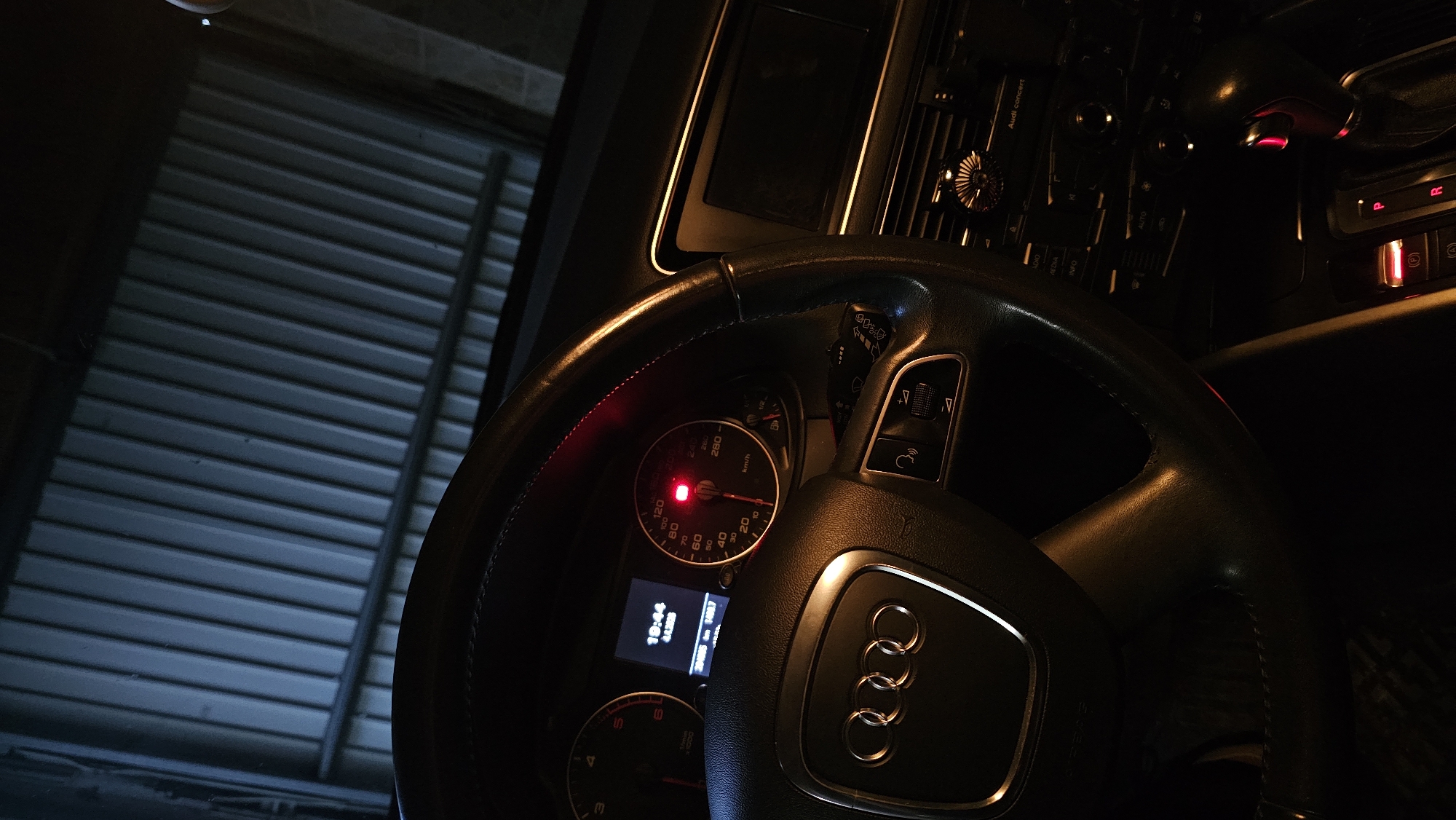 The image shows the interior of an Audi car, focusing on the steering wheel, dashboard, and a glimpse of the exterior through the front window. The steering wheel is black leather with the Audi logo prominently displayed. The dashboard features gauges, including a speedometer with a red warning light illuminated, and a digital display showing the time (19:44) and date (4.4.2026). Buttons for various car functions are visible on the dashboard. Through the front window, a slatted garage door is seen, suggesting the car is parked in a garage. There is no indication of the city of Rogachevo, Bulgaria in the image.