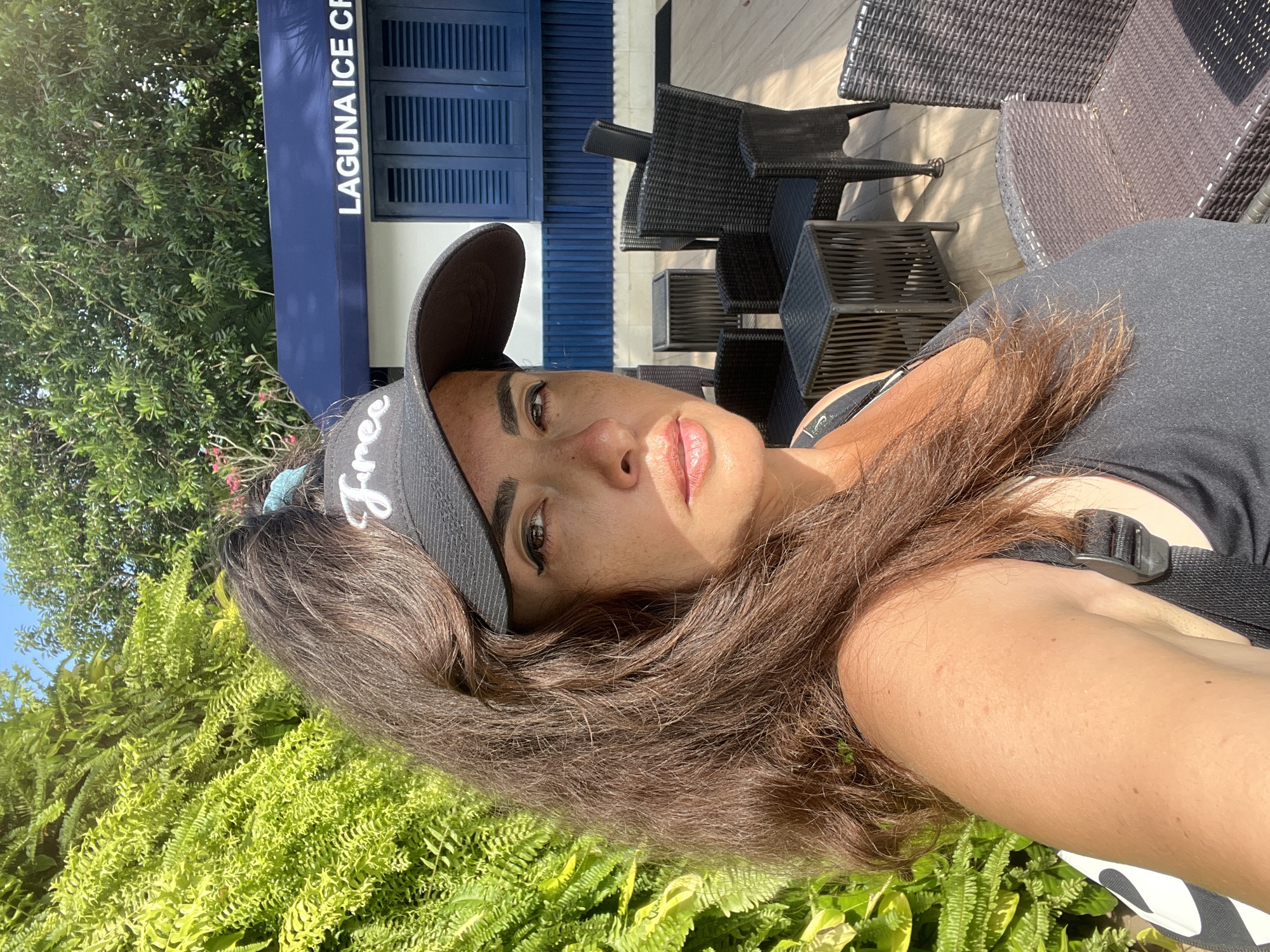 A woman is taking a selfie in front of a building that says "LAGUNA ICE CR". She is wearing a black baseball cap with the word "Free" written on it in white. She has long, brown hair that is pulled back into a ponytail. She is also wearing a black tank top. The background shows a building with blue shutters, outdoor seating with wicker chairs and tables, and lush green foliage. The scene appears to be outdoors in a sunny climate.