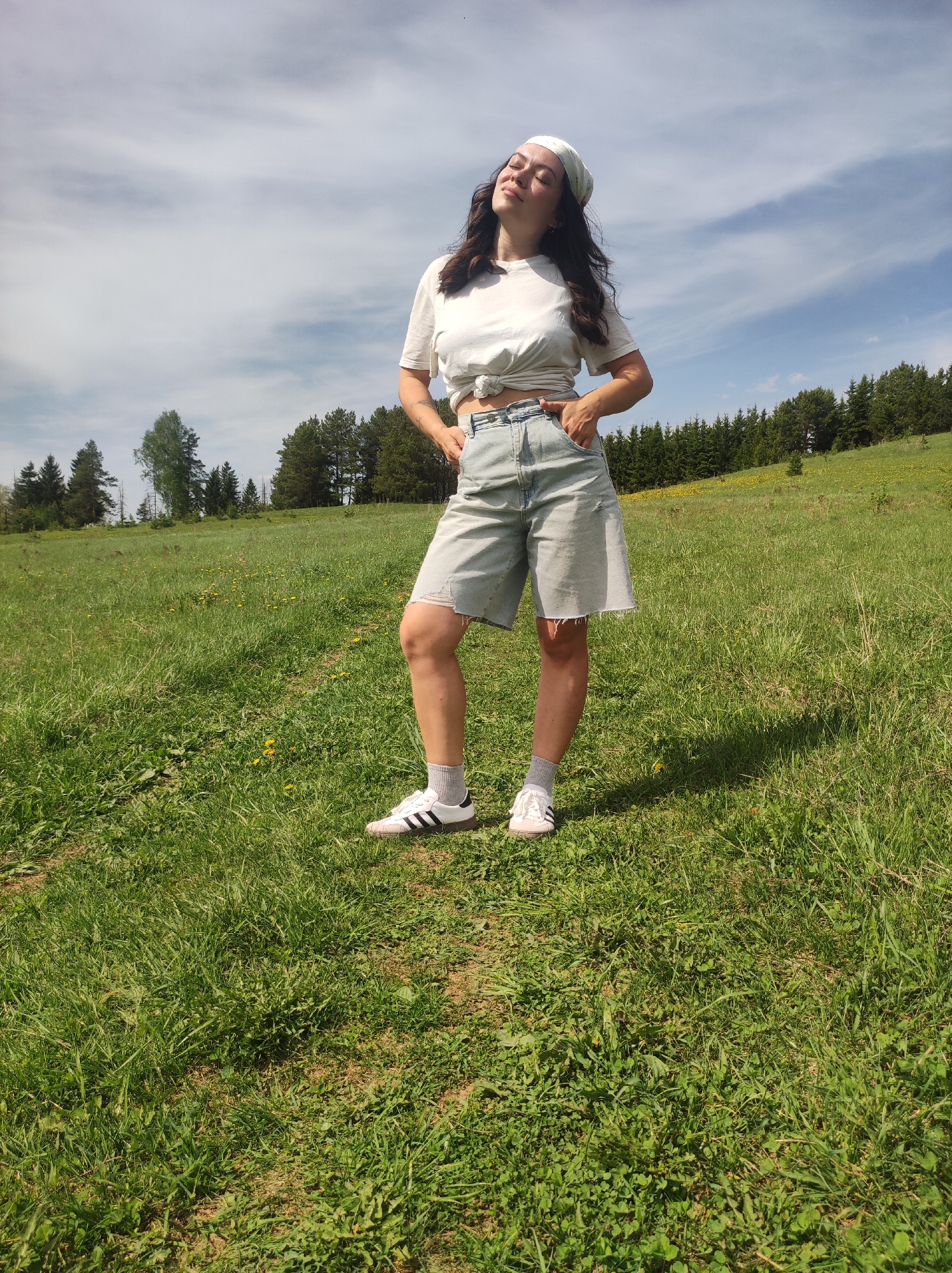 A woman stands in a grassy field on a partly cloudy day. She is wearing a white t-shirt tied in a knot at her waist, light blue denim shorts, white socks, and white sneakers with black stripes. She has a white bandana on her head and is standing with her hands in her pockets, eyes closed, and face tilted toward the sun. In the background are trees and a grassy hill. The location is consistent with a rural area near Фоки, Russia.