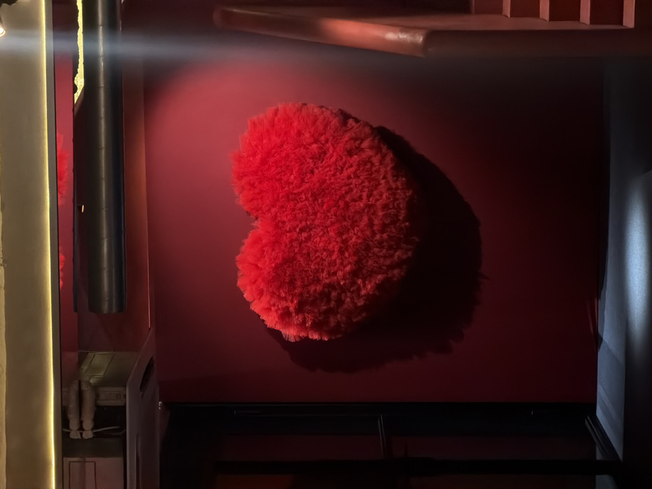 A red, fluffy heart is mounted on a dark red wall. It casts a shadow to the right. The wall appears to be part of a shelf or cabinet unit, with glimpses of a dark shelf below the heart and a ledge above. To the left, a shiny, metallic cylindrical object is visible, alongside a vertical strip of off-white wall. To the right, a bright vertical strip suggests a doorway or opening. The image is dimly lit, with light sources causing strong shadows.