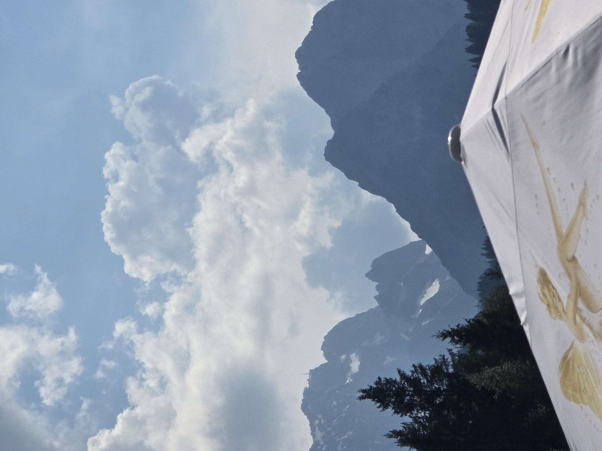 The image captures a dramatic mountain landscape under a partly cloudy sky. In the foreground, a portion of a white umbrella with a golden design is visible, tilted diagonally. The umbrella is a prominent object in the frame, partially obscuring the view of the scene behind it.
The backdrop is dominated by rugged, dark-hued mountains that rise steeply into the sky. Patches of snow are visible on some of the higher slopes, suggesting a cooler climate or season. The sky itself is a mix of light blue and soft white clouds, indicating fair weather, possibly mid-morning or afternoon.
The overall impression is one of natural grandeur and a serene outdoor setting. The steepness of the mountains and the vastness of the sky create a sense of awe. The presence of the umbrella suggests a person or people are present, perhaps enjoying the view or seeking shade from the sun. There are no visible people or text in the image. The location context of Garmisch-Partenkirchen, Germany, further reinforces the interpretation of a mountainous alpine environment.