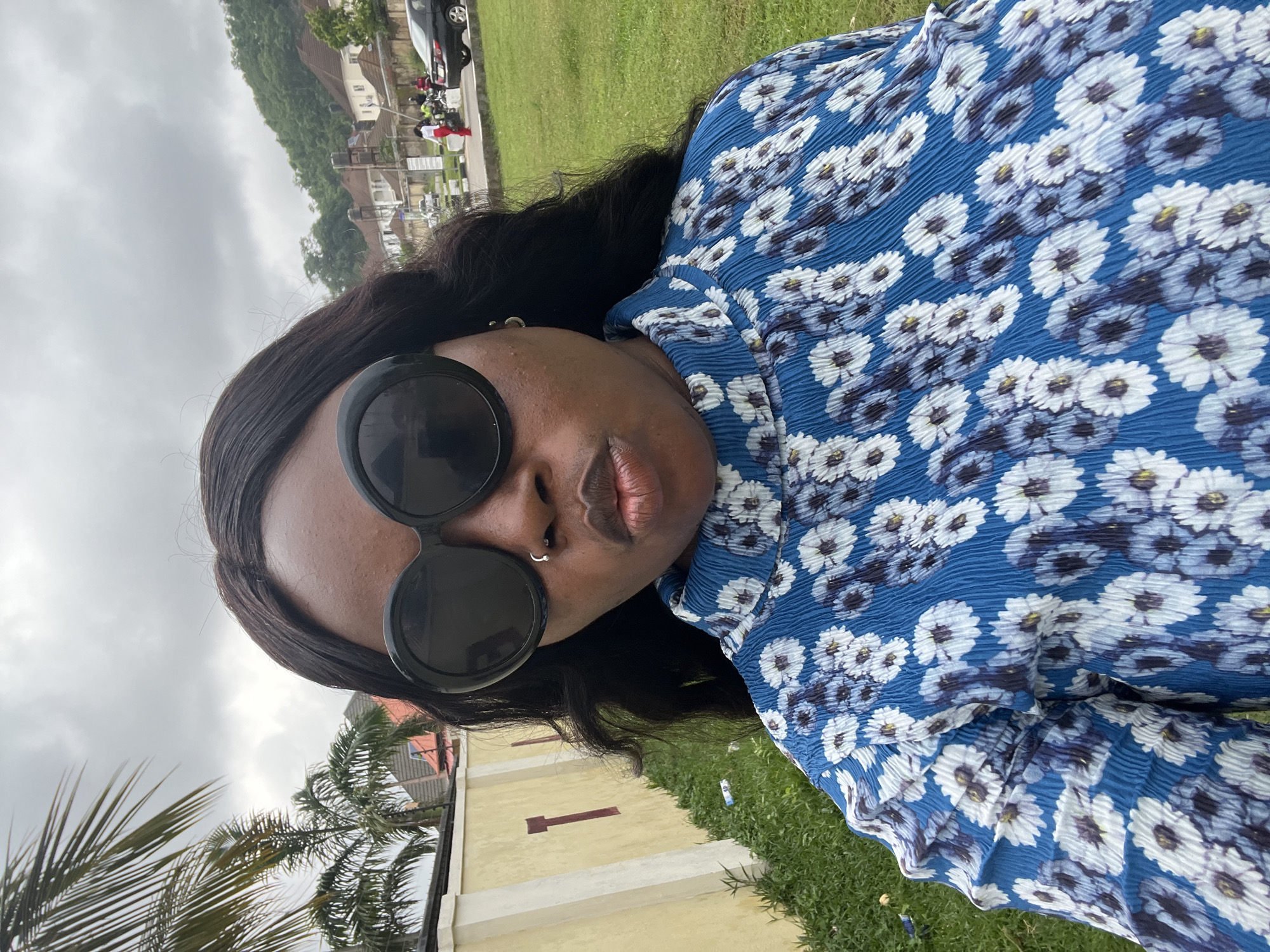 A medium shot captures a Black woman wearing large, circular black sunglasses and a blue top with a white floral print. She is looking forward and has her lips pursed, as if posing for a selfie. The background shows a suburban area with houses and lush green trees, suggesting a location in Abuja, Nigeria. A yellow wall with red vertical markings runs along the left side of the frame, with some overgrown grass and debris at its base. A paved road is visible in the distance, with a few people and vehicles, including a black car and motorcycles. The sky is overcast, indicating a cloudy day.