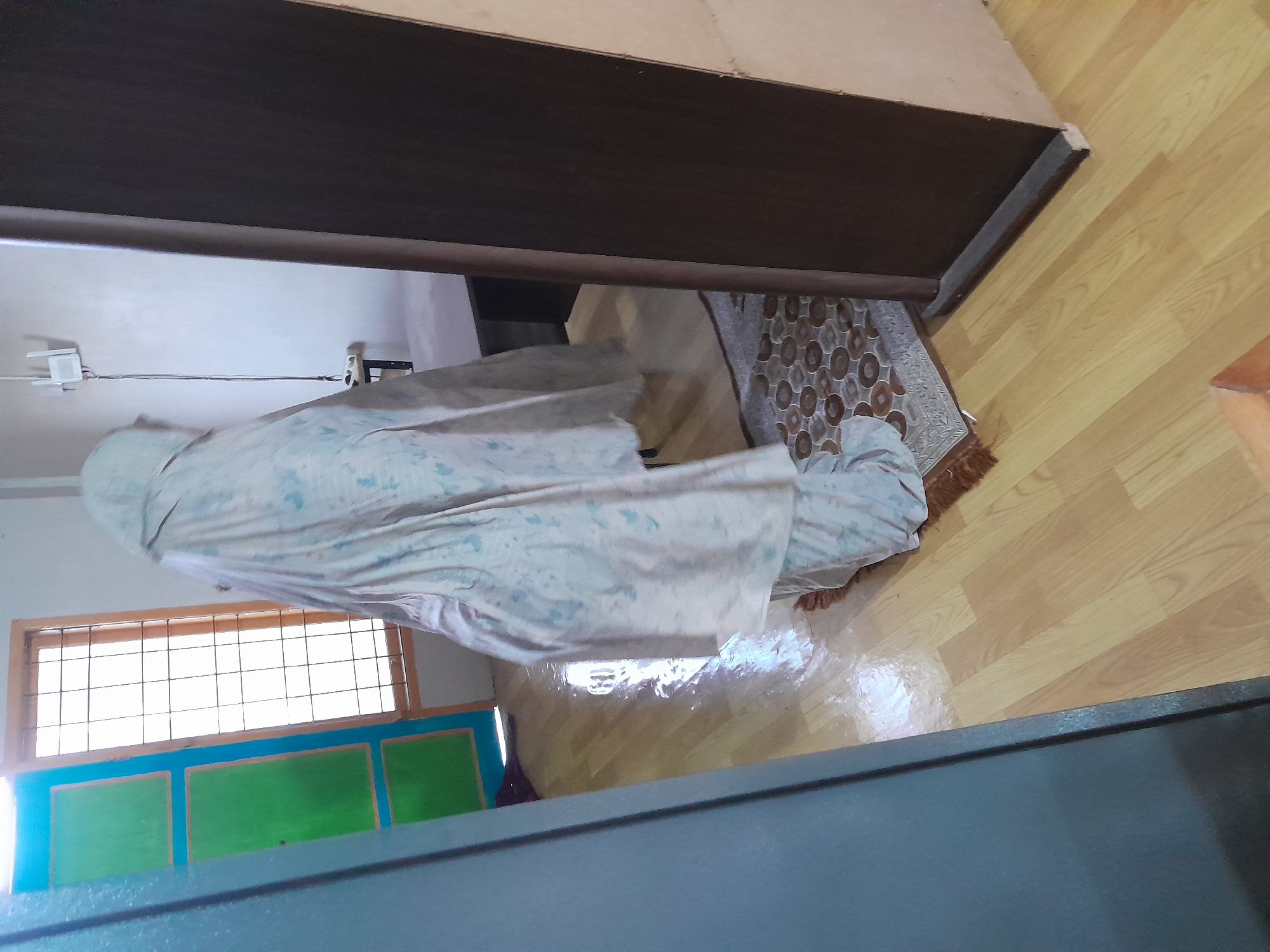 A person covered in a light-colored, patterned sheet is kneeling on a patterned rug in a room with light-colored wooden floors. A window with a wood frame and metal bars is visible on the left. The lower portion of the window is painted blue and green. A brown wooden beam is present in the upper part of the image. The location is Tingkeummanyang, Indonesia.