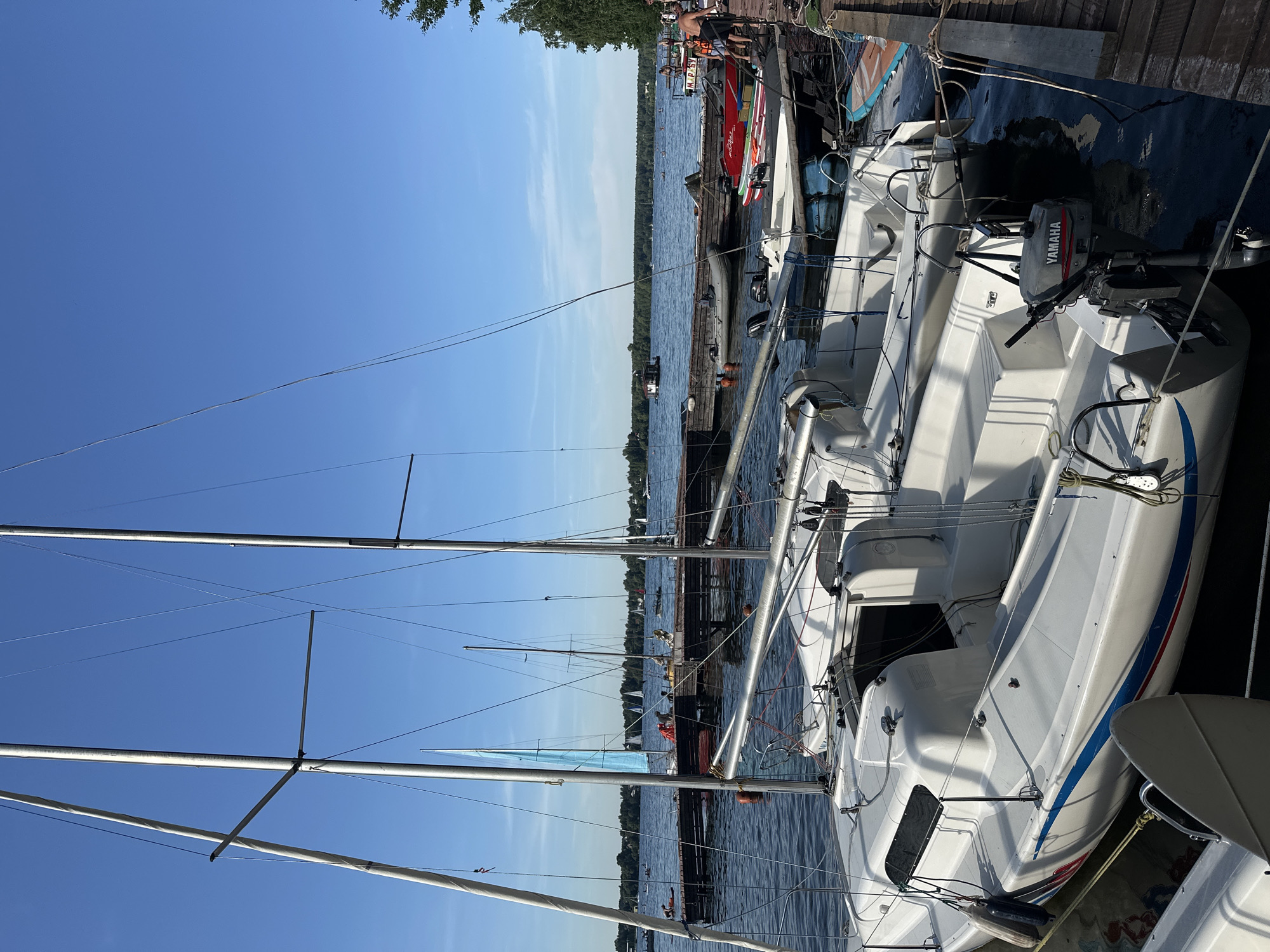 This is a medium-shot, outdoor, eye-level, horizontal photograph of a marina on a sunny day, likely at the Tourist Resort "Klyazminskoe Reservoir" in Russia. The foreground is dominated by the white hulls of two docked sailboats, their masts and rigging silhouetted against a clear blue sky. One of the sailboats has a visible Yamaha outboard motor mounted on its stern. The boats are secured to a wooden dock, which extends into the water and is also visible in the mid-ground.
Beyond the sailboats, a wooden pier is filled with activity. Several small boats are moored there, including a gray inflatable dinghy and a red and white rigid inflatable boat. People are gathered on the pier, some appearing to be preparing boats, while others are socializing or observing the water. Further out on the reservoir, other sailboats are visible, some with sails up, indicating ongoing activities or races. The far shore is lined with a dense forest, and a few buildings or structures can be seen among the trees. The water is calm, with gentle ripples reflecting the clear sky. The overall scene depicts a leisurely summer day at a waterside recreational area.