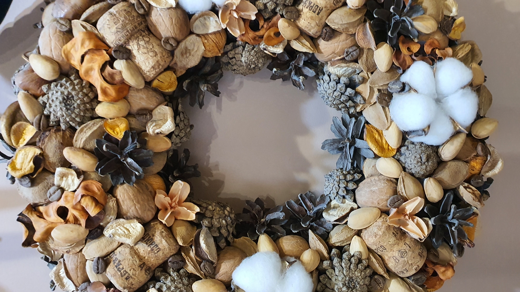 This media file displays a close-up, overhead view of a decorative wreath, likely intended for autumnal or holiday display. The wreath is circular and appears to be made from a variety of natural elements, including nuts, dried leaves, pinecones, and coffee beans. Notable objects within the wreath include whole walnuts, almonds, and what appear to be pieces of cork. There are also some small, dried floral elements in shades of beige and orange, and two white cotton bolls. The overall color palette is earthy and muted, with browns, beiges, and grays dominating.
The wreath is arranged densely, creating a textured and rustic appearance. The subjects are purely objects, with no people or animals visible. The scene is static, with no activity or event taking place beyond the display of the wreath itself.
There is some visible text on the cork pieces, though it is partially obscured and difficult to read clearly in the provided image. The background is a plain, light-colored surface, suggesting an indoor setting. There are no cues regarding time of day or weather, and no discernible emotions are conveyed by the inanimate objects. The location context of Krasnoyarsk, Russia, does not provide any direct visual cues within the image itself.