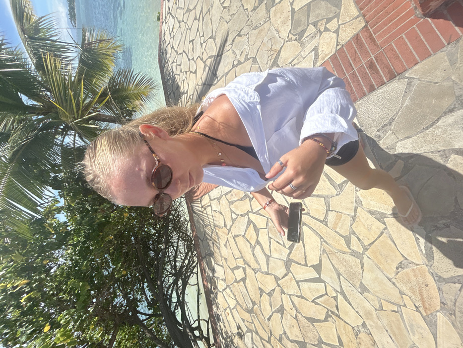 An outdoor scene in Sainte-Anne, France, features a female individual on a sunlit, irregularly-paved stone pathway. The subject, with blonde hair, wears tortoiseshell-patterned oval sunglasses, a white button-up shirt unbuttoned over a black bikini top, black shorts, and white flip-flops. Her attire is accessorized with multiple rings on both hands, several beaded bracelets on both wrists, and a layered necklace with small pendants. She holds a dark-colored smartphone horizontally in her left hand, positioned low, while her right hand is near her chest. The pathway's irregular light-colored flagstones transition to a reddish-brown brick structure on the right side. The background shows lush green tropical foliage, including palm trees, against a clear, turquoise body of water under a bright blue sky.