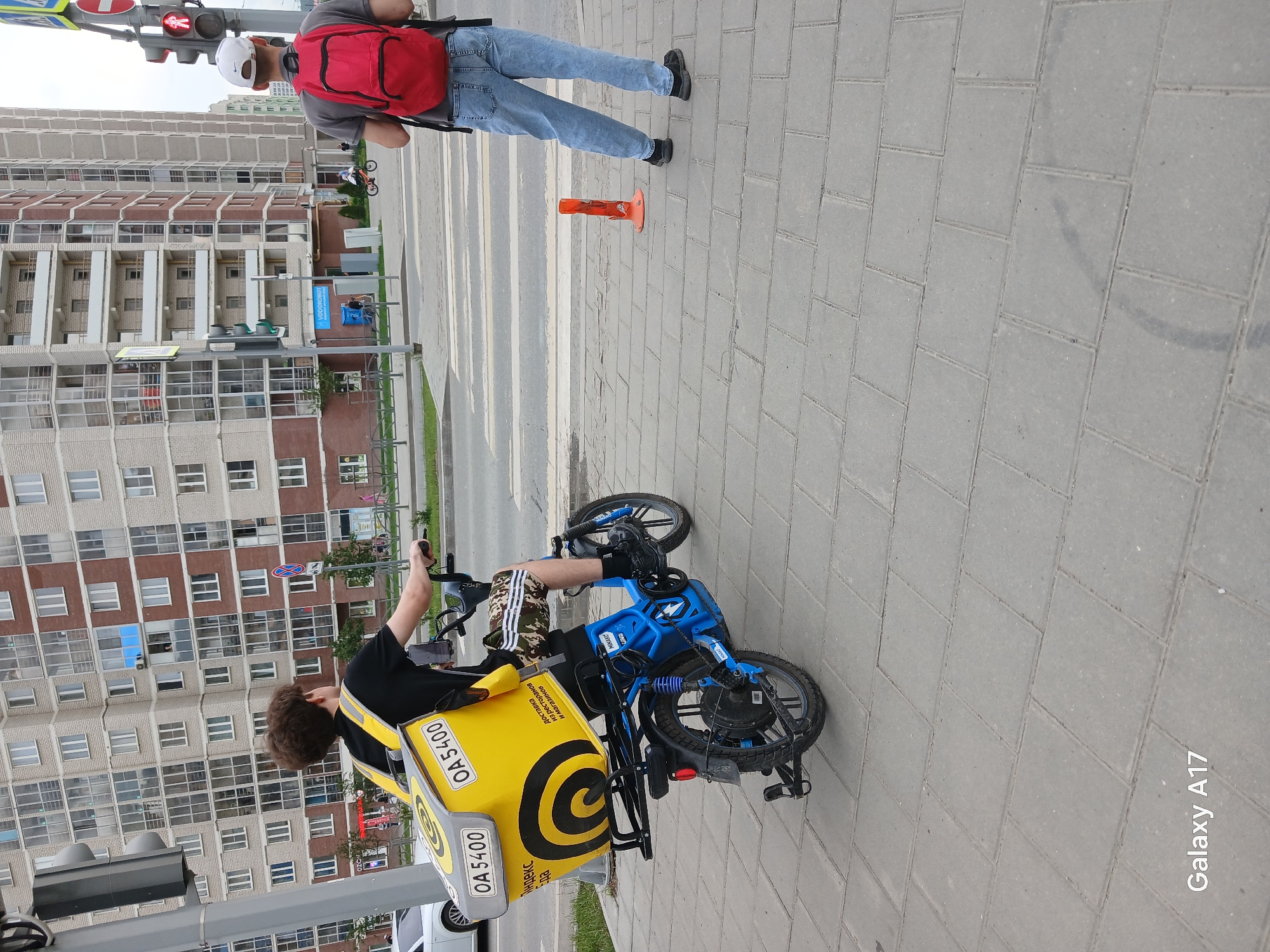 An individual wearing a black t-shirt, camouflage shorts, and black socks is riding a blue electric scooter. The scooter has a large yellow delivery box mounted on the rear, with "OA 5400" and Russian text indicating delivery services. The rider is positioned on a paved sidewalk next to a crosswalk. In the background, a multi-story apartment building with numerous windows and balconies is visible. Another person, wearing a grey t-shirt, blue jeans, a white baseball cap, and a red backpack, stands on the opposite side of the crosswalk, facing away from the camera. A red traffic cone is positioned on the sidewalk near this individual. The scene appears to be a street in Yekaterinburg, Russia, evidenced by the text on the delivery box, the architectural style of the building, and the "Galaxy A17" watermark.
