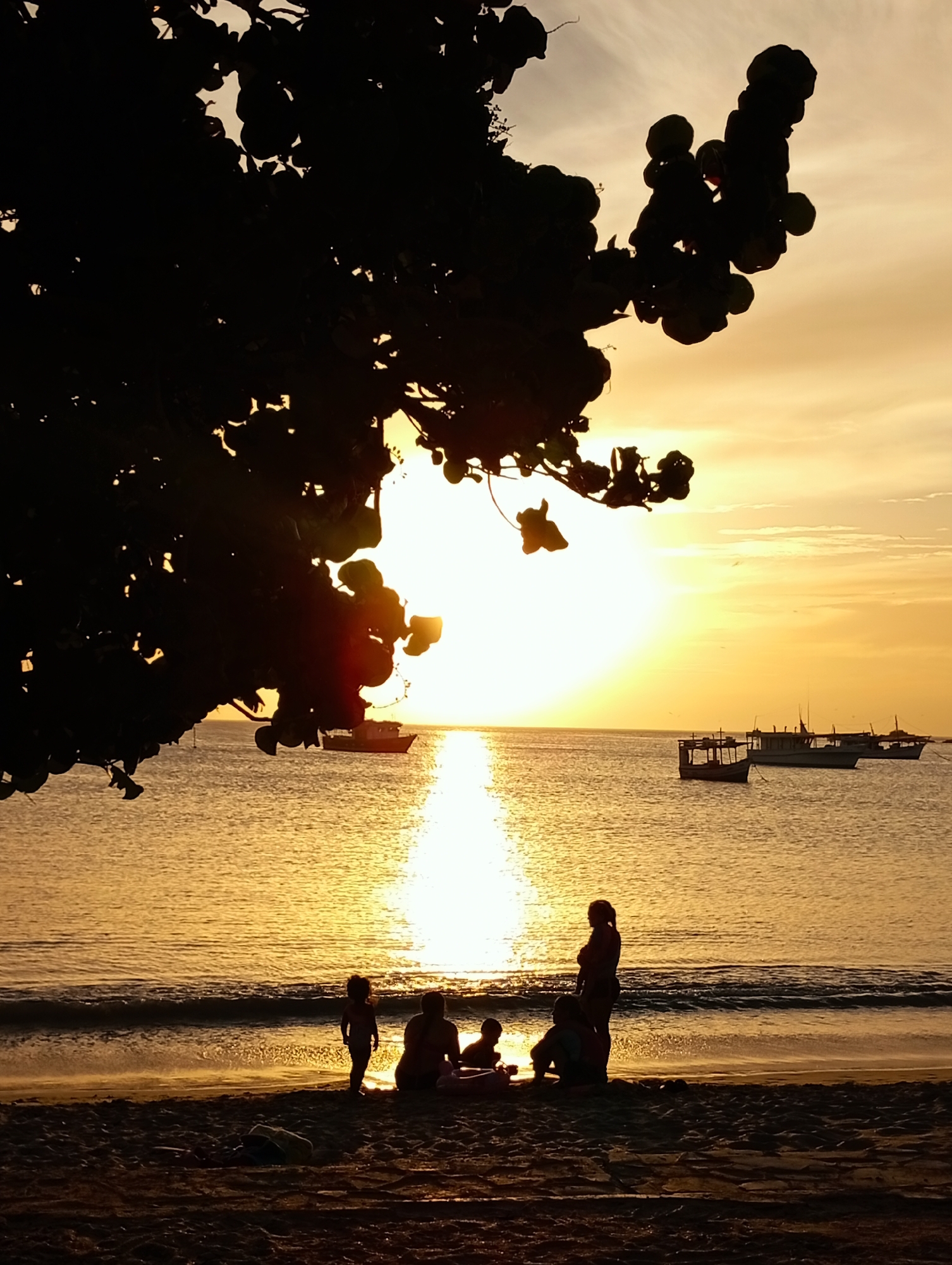 The image shows a beach at sunset in Juan Griego, Venezuela. Silhouetted in the foreground are a group of people, including children and adults, sitting and standing on the sand. In the water are several boats. A silhouetted tree branch and leaves are in the upper left of the image. The sun is setting over the ocean.