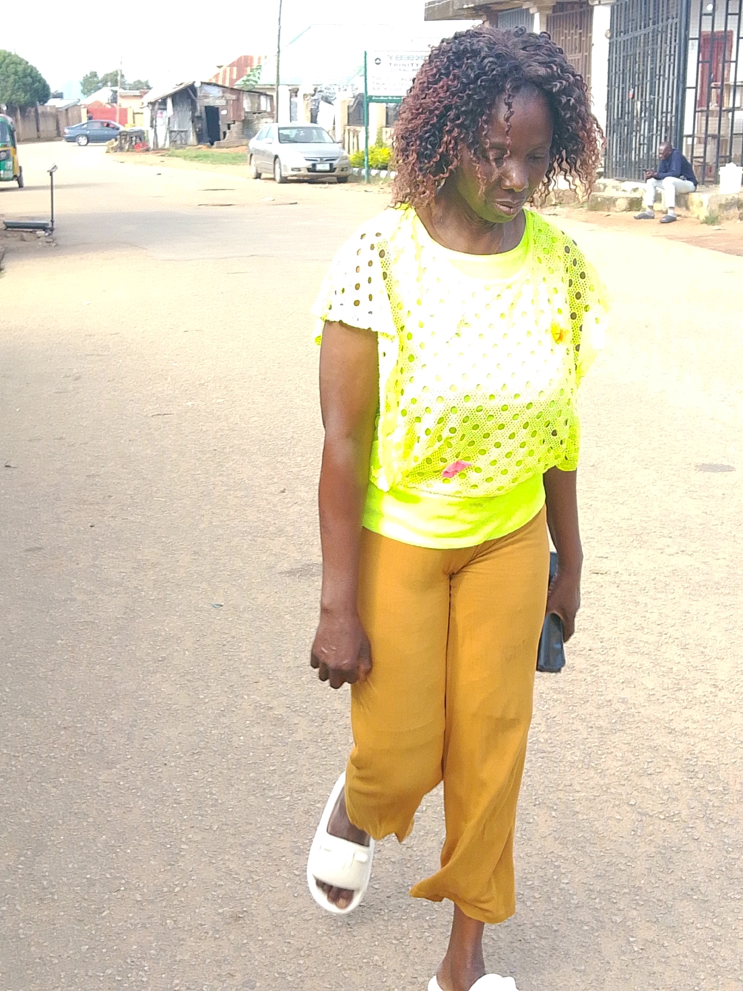 A woman with curly brown hair walks on a paved road in Jos, Nigeria. She is wearing a yellow, perforated mesh top over a neon yellow undershirt and loose-fitting mustard-yellow pants. Her left foot is adorned with a white slide sandal, and she is carrying a dark-colored phone in her right hand. The environment appears to be a street with buildings lining the sides, some with corrugated metal roofs. A few cars are visible in the background, including a silver sedan and a blue car. The sky is overcast.