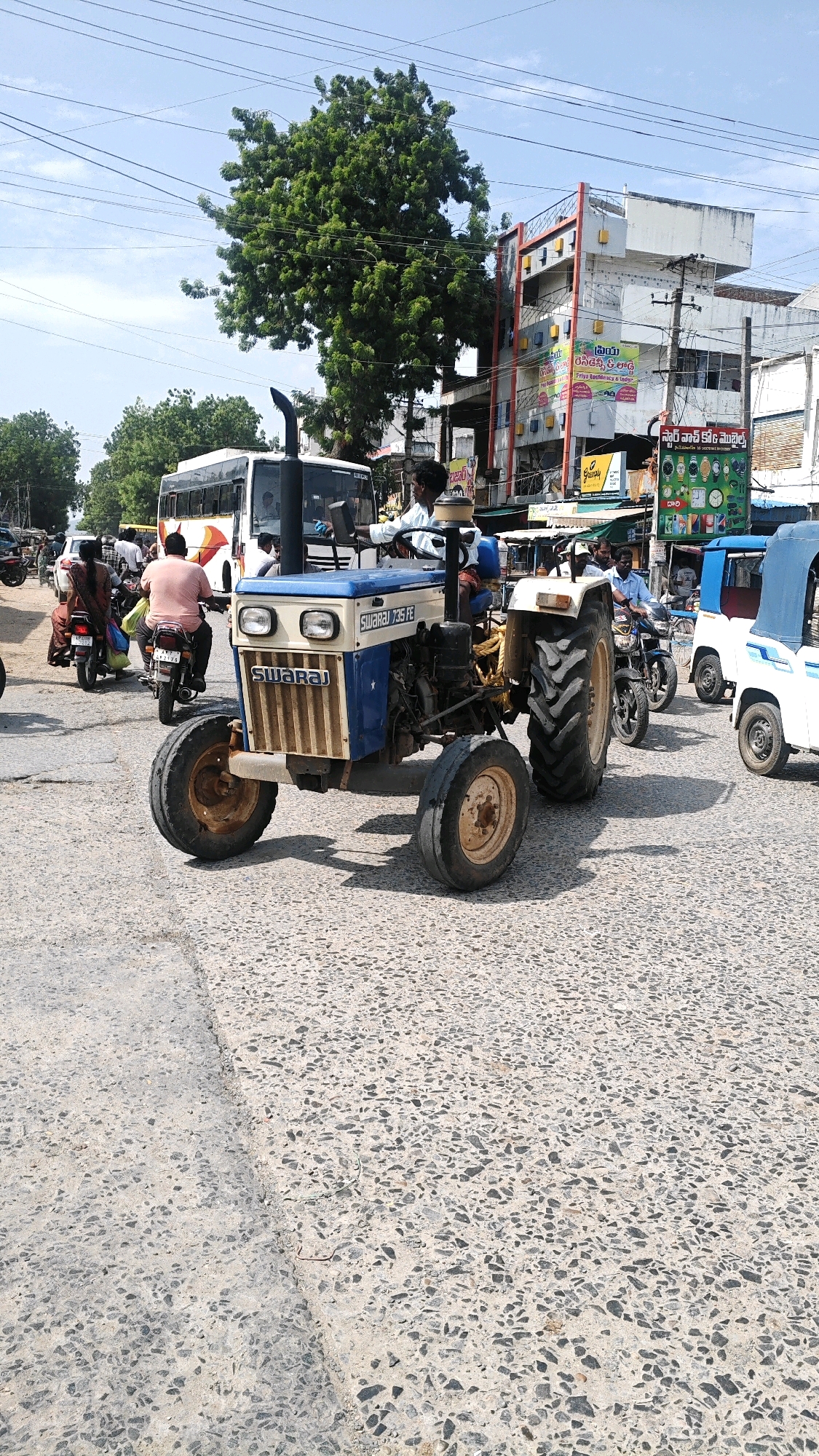 In Pamur, India, on a sunny day, a blue and beige Swaraj tractor is in the foreground of a street scene. The tractor is driven by a man wearing a white shirt. Behind the tractor, a white bus with a red and orange stripe travels down the road. Further in the background, several people are seen on motorcycles and a white auto-rickshaw with a blue canopy is also visible. The street is paved with concrete and lined with buildings on the right side. A large green tree stands prominently on the left side of the image. Overhead, power lines crisscross the sky.