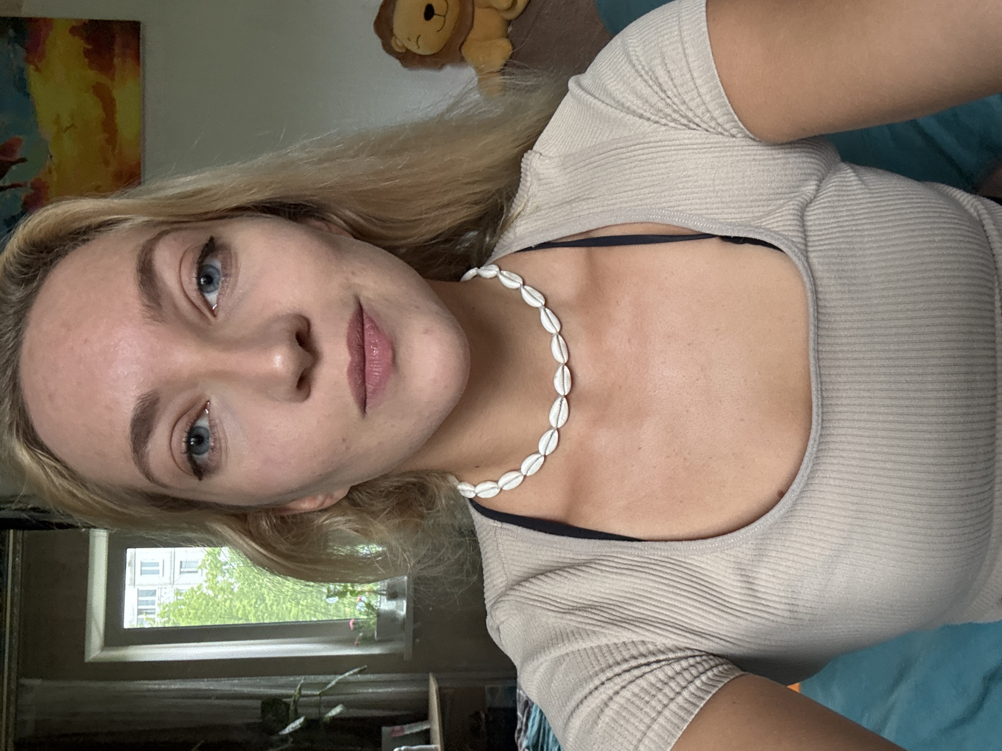 A woman with blonde hair and blue eyes is depicted from a high-angle perspective. She is wearing a light-colored ribbed top and a necklace of white shells. The background includes a window with a view of greenery and buildings, suggesting an urban setting. Other objects visible include a stuffed lion toy and a painting. It is not possible to determine if this is Minsk, Belarus.