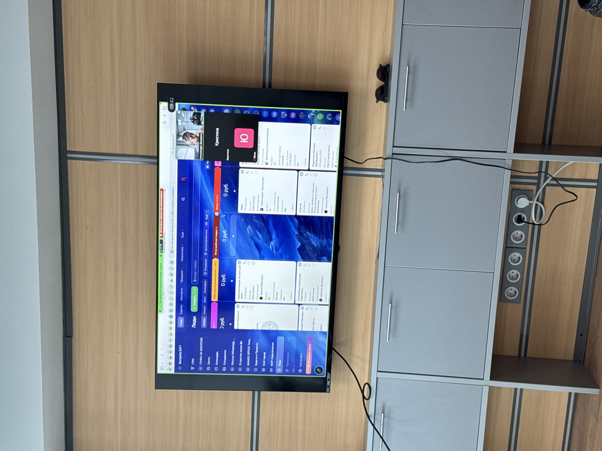 The image shows a horizontally oriented view of a flat-screen television mounted on a wall with light-colored, horizontally striped panels. The screen displays a computer interface, possibly a CRM or project management software. The interface features a list of contacts or leads, with a prominent section displaying profile information and contact details, including names like "Кристина Соломеннкова" and "Александр Черноусова." A smaller window on the top left shows a video call with a person.
To the right of the television is a grey cabinet with silver handles and visible electrical outlets. The environment appears to be an office, indicated by the wall paneling and cabinet. Sunglasses are placed on top of the cabinet.
There is no specific visual information present that indicates the location is in Perm, Russia.
