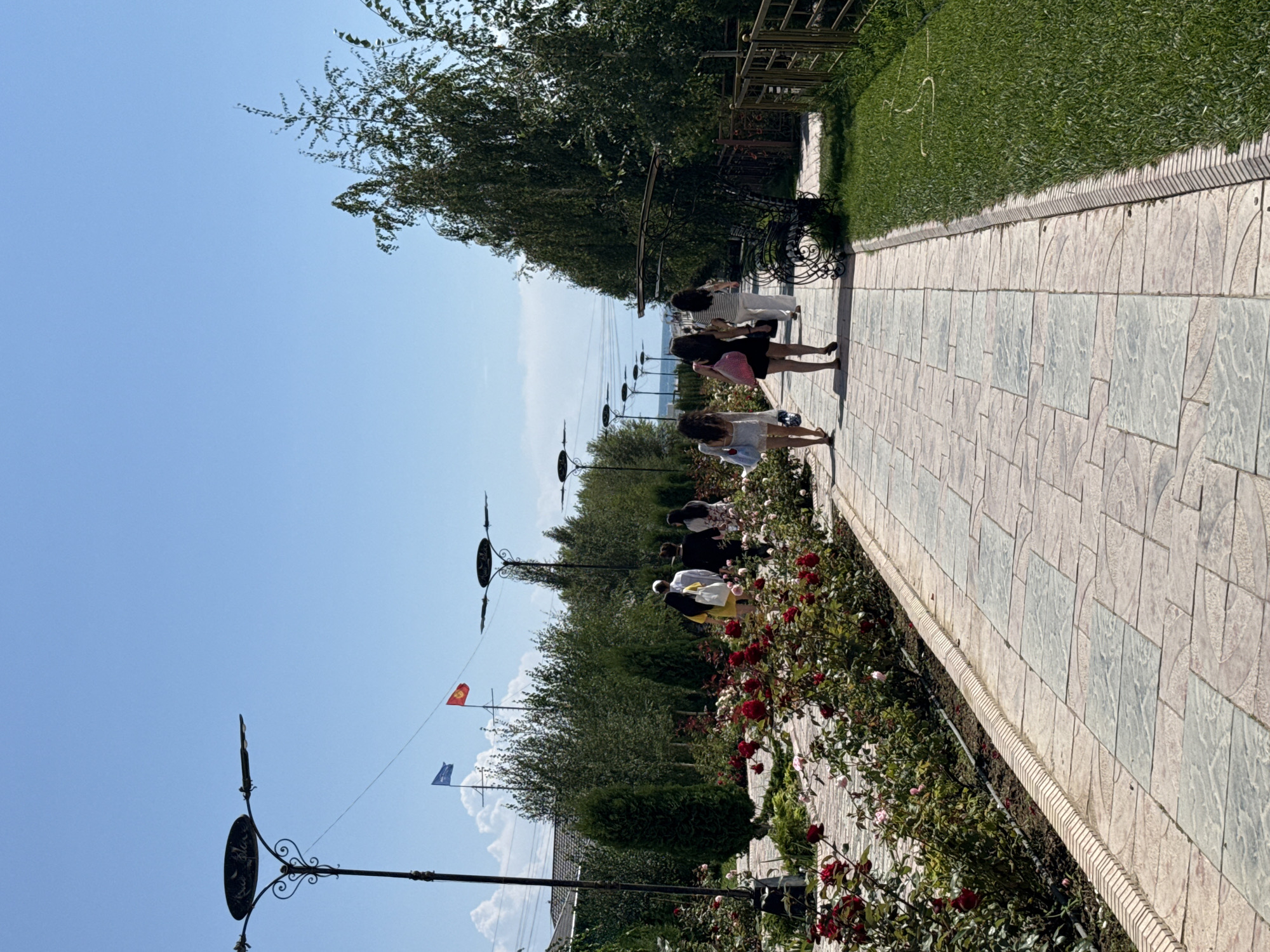 In Sary-Oy, Kyrgyzstan, on a paved pathway, a group of individuals are walking, some carrying bags. The pathway is lined with a vibrant rose garden to the left and a well-maintained lawn to the right, bordered by a fence. The sky above is clear and blue, with two flags visible in the distance. The scene suggests a leisurely stroll through a park or resort area.