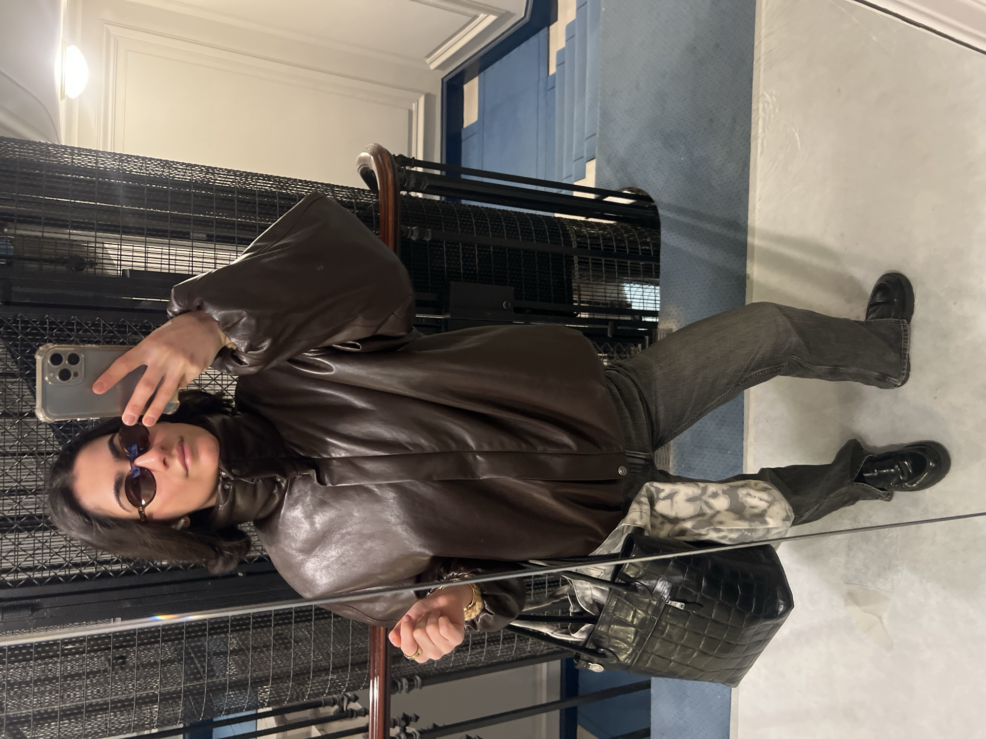 A woman is taking a selfie in an elevator in Paris, France. She is wearing sunglasses, a brown leather jacket, dark-colored jeans, and black shoes. She is holding a black leather bag. The elevator has a black metal cage design. The background shows a hallway with blue carpet and white walls. There is a light fixture on the wall above the elevator.