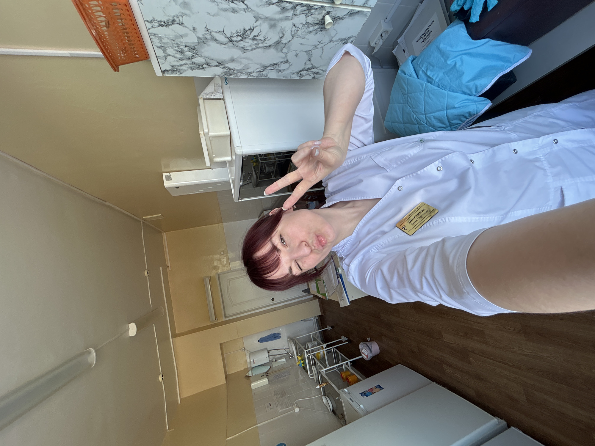 A woman in a white medical coat, likely in her 30s, poses for a selfie in what appears to be a medical office or clinic in Фоки, Russia. She is making a peace sign with her right hand. She has red hair and is looking directly at the camera with a playful expression. The background includes medical equipment, white cabinets, and tiled floors, suggesting a sterile environment. A name badge is visible on her coat.