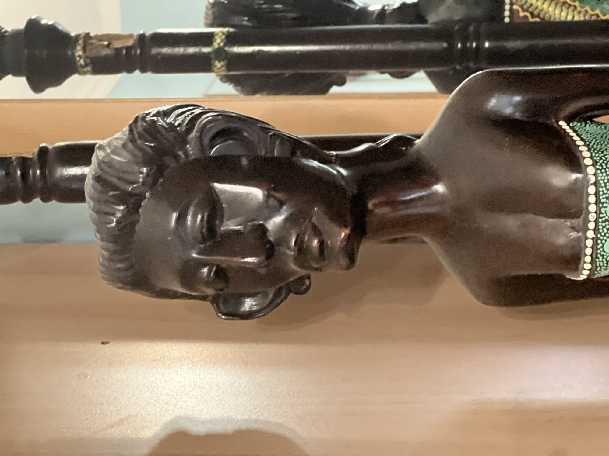 A dark brown, polished wooden sculpture of a woman is positioned in the foreground, facing slightly left. The sculpture depicts the upper torso and head of the woman. She has distinct facial features, including a narrow nose, full lips, and large ears that extend outwards. Her hair is styled in waves around her head, and her eyes are closed or downcast. The sculpture is adorned with a beaded necklace or garment around her chest, featuring a pattern of small white dots on a green background.
To the left of the sculpture is a plain, light brown wall. To the right of the sculpture, a dark brown, ornate wooden pillar stands, likely part of a piece of furniture or a decorative stand. In the background, a reflection is visible in what appears to be a mirror, showing another dark wooden pillar and a hint of a light blue wall. The overall environment appears to be indoors, possibly a home or a gallery, with soft lighting. The scene is static, with no apparent action or interaction occurring.