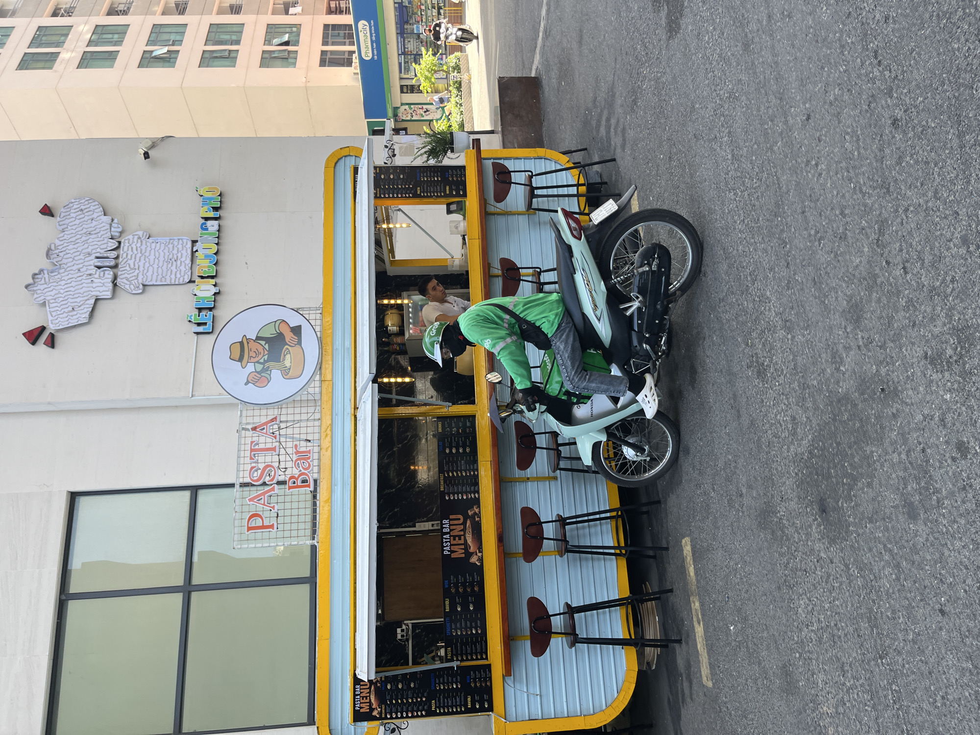 The image shows a restaurant called "PASTA Bar", located in the city of Bac Nha Trang Ward, Vietnam. The restaurant has an outdoor counter with bar stools. A delivery driver on a moped is parked next to the restaurant, presumably waiting to pick up an order. The restaurant's name and menu are clearly visible. There are buildings visible in the background. A pharmacy can also be seen in the background.