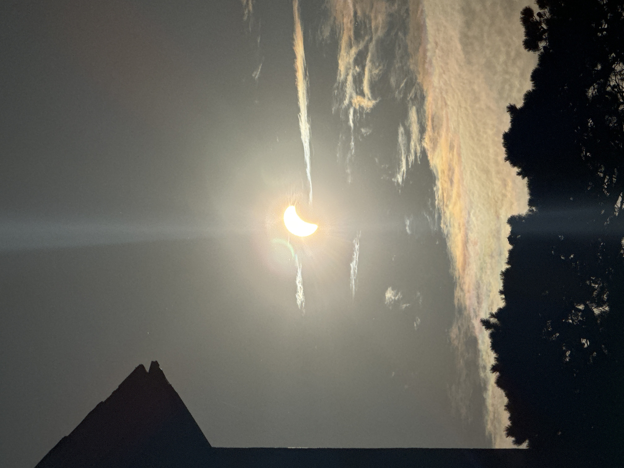 The image depicts a partial solar eclipse observed from what appears to be a residential setting in Waterford, Ireland. In the foreground, a dark silhouette of what could be a roofline from a building is visible on the left side of the frame, and a dense silhouette of evergreen trees forms the bottom portion of the image. The sky is overcast with wispy clouds, some of which are illuminated with iridescent colors, suggesting the sun's rays are refracting. The partial solar eclipse is the focal point, appearing as a bright crescent of light in the upper central part of the sky. No people are visible in the image. The overall scene captures a natural celestial event occurring within a suburban or semi-rural environment.
