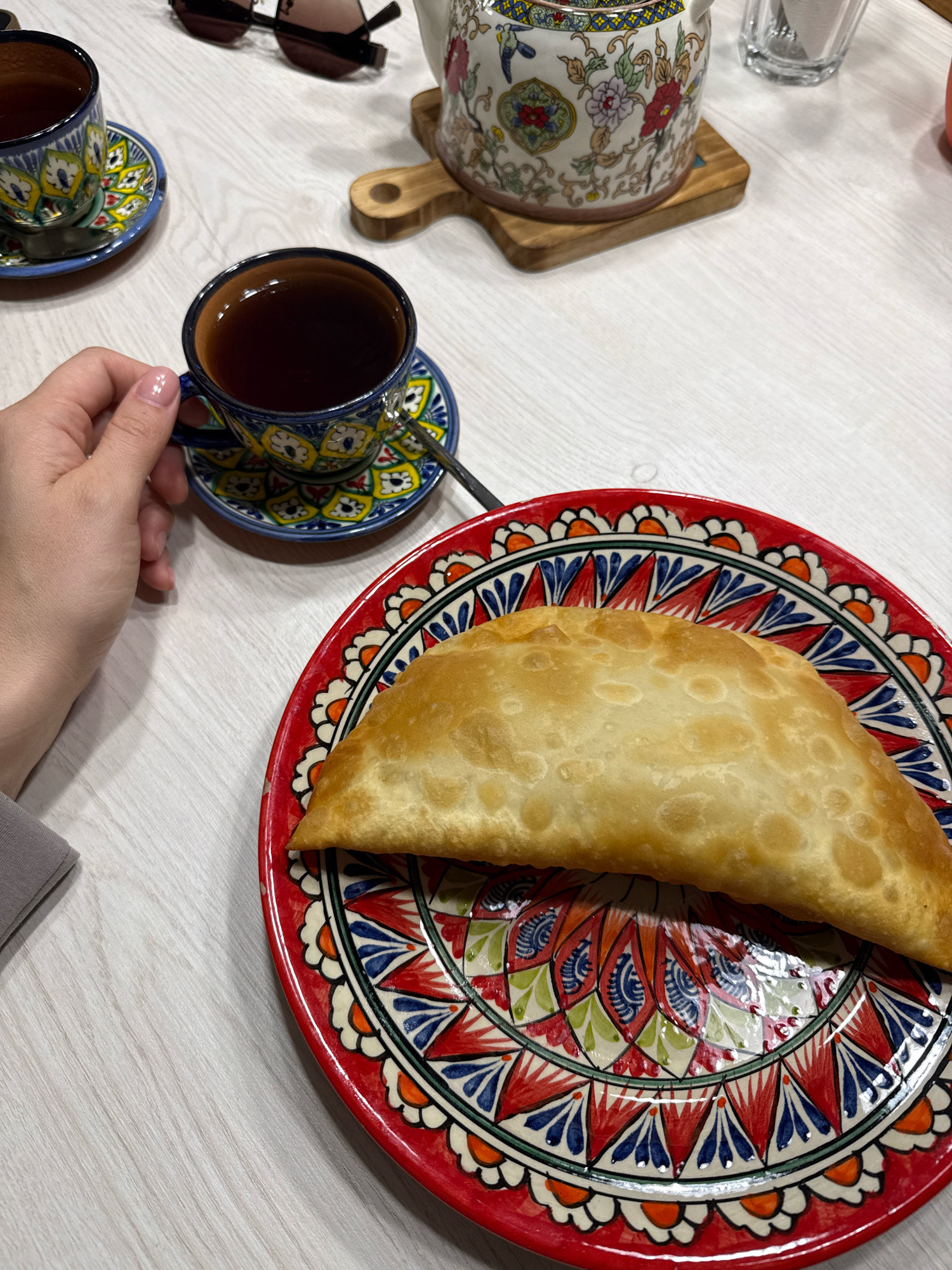 A close-up, high-angle shot shows a person's hand holding a cup of dark tea over a decorative saucer. Next to it, a plate with a golden-brown pastry sits on a white wooden table. In the background, a teapot with floral patterns and a pair of sunglasses are visible. The setting appears to be a home or cafe in Chaikovsky, Russia, based on the visual cues of the tea set and pastry which are common in Russian cuisine.