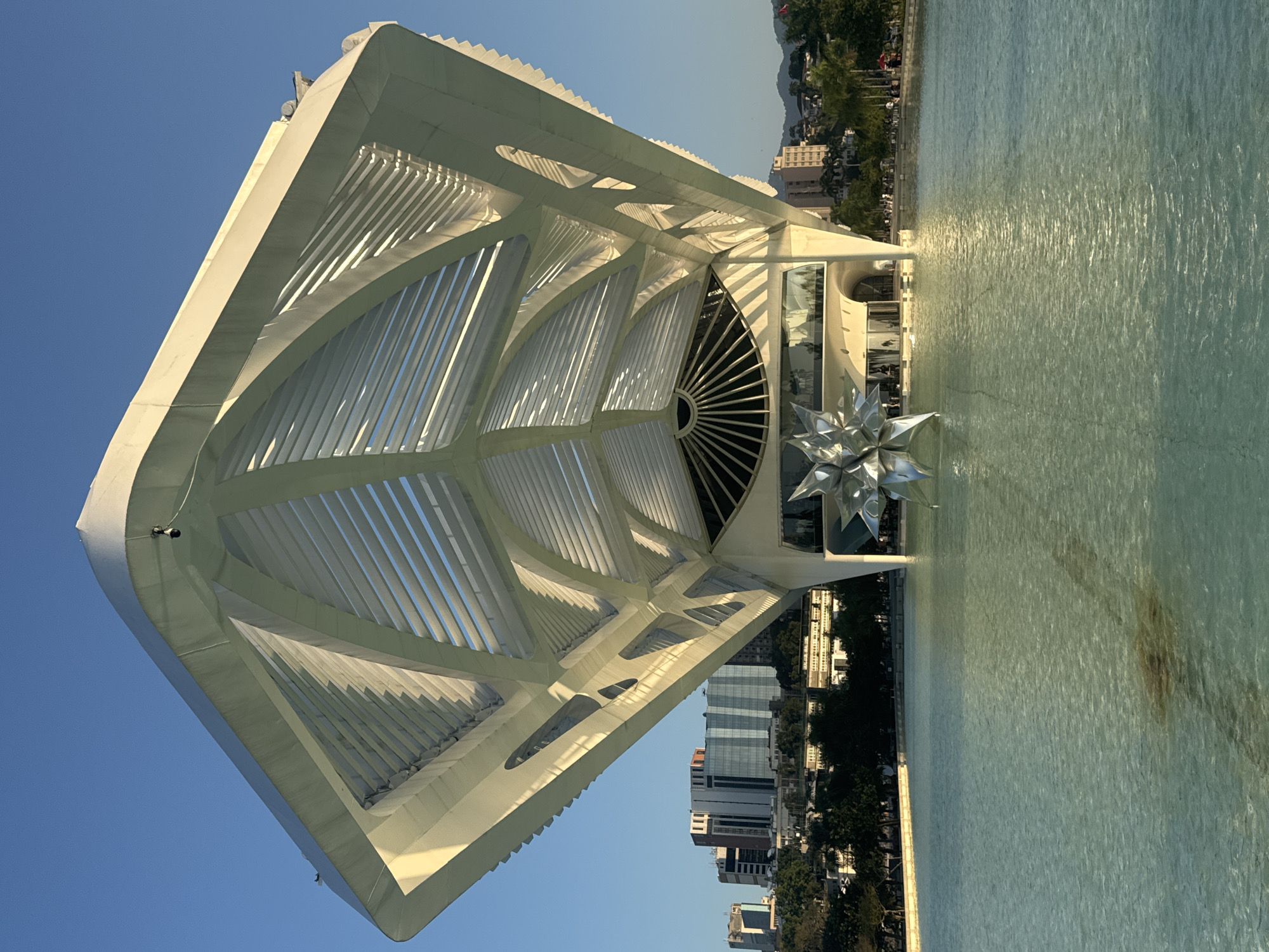 The image depicts the Museum of Tomorrow (Museu do Amanhã) in Rio de Janeiro, Brazil, a striking example of modern architecture. The museum's distinctive design, resembling a large white wing or boat, is prominently featured. It is situated beside a tranquil body of water, likely the Guanabara Bay, which reflects the clear blue sky and the building's structure. The water appears calm with subtle ripples, suggesting a peaceful day.
The architecture of the museum is characterized by its large, cantilevered roof with a complex system of white fins or louvers, creating a dynamic interplay of light and shadow. These elements extend outwards, giving the structure a futuristic and organic appearance. In front of the museum, a large, silver, star-shaped sculpture is visible, adding an artistic focal point.
In the background, the cityscape of Rio de Janeiro unfolds, with buildings and lush green trees lining the waterfront. The presence of these urban elements contrasts with the serene water and the avant-garde design of the museum. The lighting suggests it is daytime, with the sun casting bright highlights on the building and casting shadows, possibly indicating late afternoon. The sky is a deep, clear blue, indicating fair weather. A few people can be seen along the waterfront, suggesting public access and activity around the museum. No visible text is present in the image. The overall impression is one of grandeur, innovation, and a harmonious blend of art, science, and the natural environment.