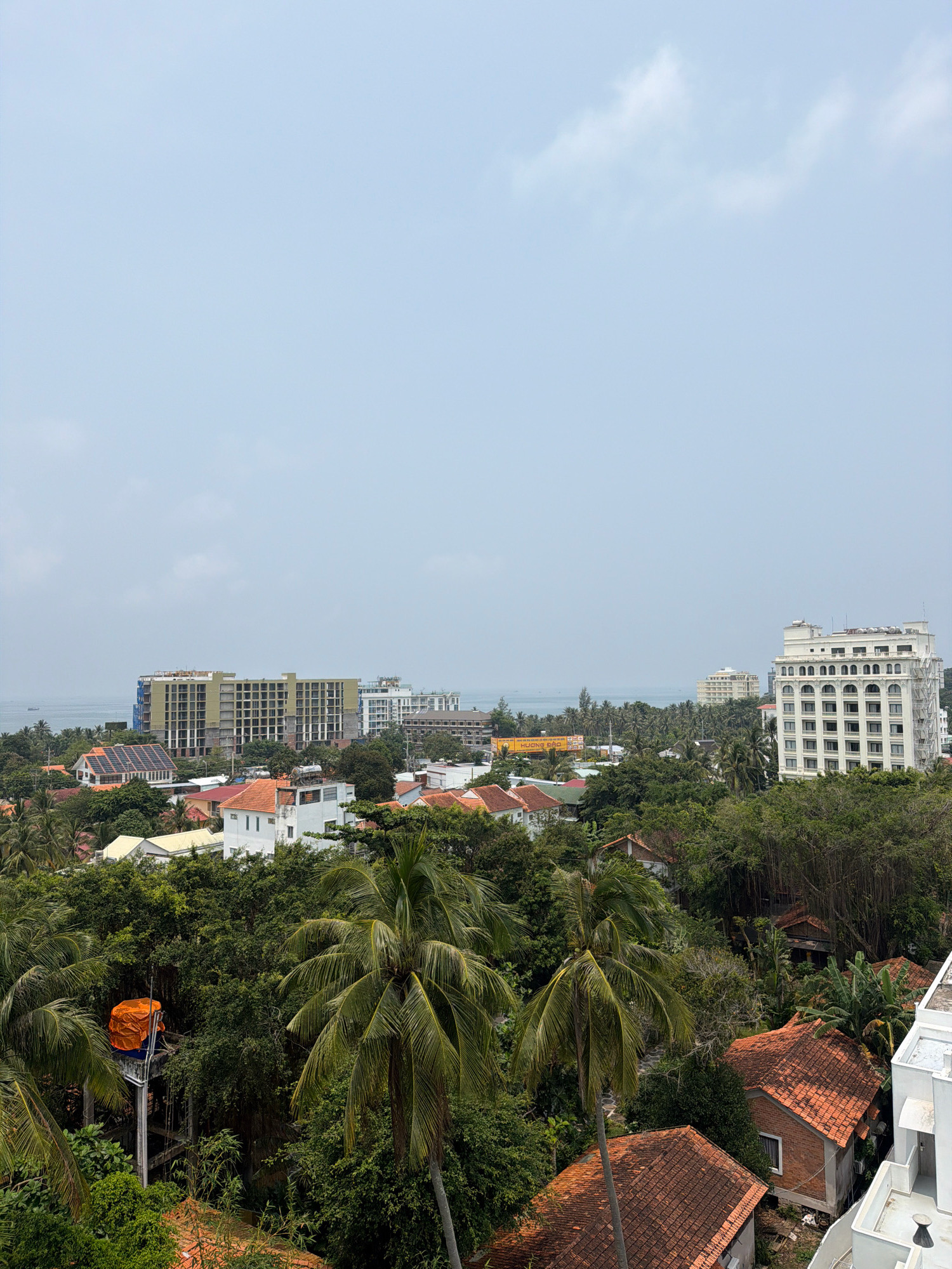 The image captures a panoramic view of a coastal city on Phú Quốc Island, Vietnam, likely during the daytime under a hazy, overcast sky. The foreground is dominated by lush green tropical foliage, including palm trees and dense bushes, interspersed with the terracotta-tiled roofs of residential houses.
Further into the scene, a cluster of buildings of varying sizes and architectural styles are visible. To the left, a large, multi-story building appears to be under construction, its exterior frame exposed and wrapped in blue netting. Adjacent to it are other residential structures with orange-tiled roofs and some with solar panels.
Dominating the right side of the mid-ground is a prominent, multi-story white building with arched windows and a distinctive facade, suggesting it might be a hotel or resort. In the distance, the calm, hazy blue waters of the sea are visible, with a faint outline of what could be a ship. Another multi-story building, lighter in color, stands further back near the coastline.
A notable object in the mid-left foreground is a tall, slender structure, possibly a water tower, topped with an orange tarp covering a large cylindrical object. The overall impression is one of a developing urban area integrated with natural tropical landscapes, situated by the sea. No people are visible in the frame.