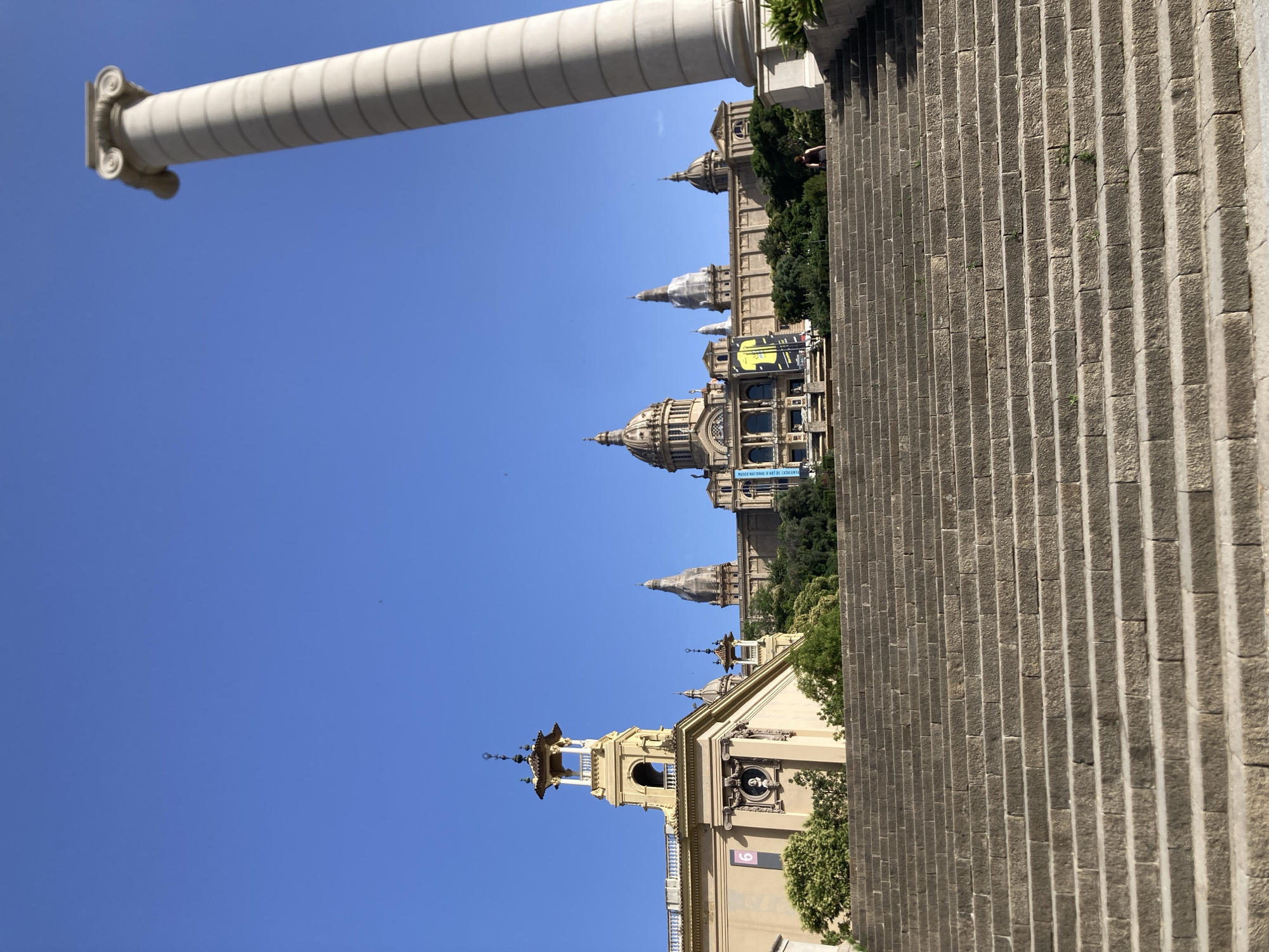 A wide stone staircase ascends towards the Palau Nacional, a large building in Barcelona, Spain, topped with multiple domes and a prominent central tower. The sky is a clear, bright blue. To the right of the staircase, a tall, white column with a scrolled capital rises into the frame. A single person is visible walking up the stairs near the top. Lush green trees and foliage surround the base of the Palau Nacional. The building displays banners, one of which is yellow and features a graphic design. Another banner on the left side of the building shows the number "6" in a pink square. The architecture of the Palau Nacional features classical elements, including columns and ornate facades.