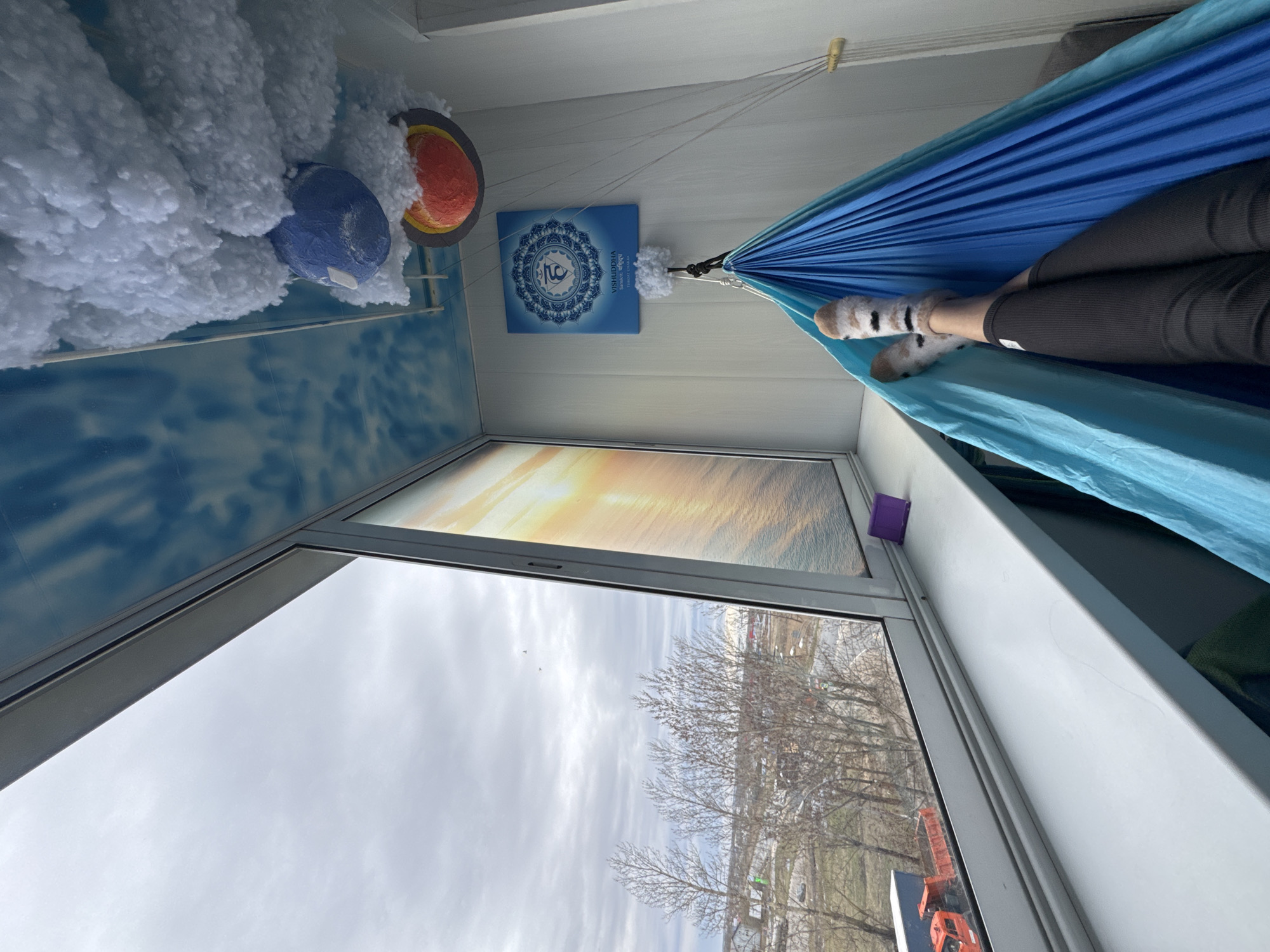 A view from inside a room looking outward through a window on what appears to be a cloudy day in Kladovitsy, Russia. Inside, a blue hammock is suspended, and a person wearing patterned socks and dark pants is reclined within it. On the wall, there's a blue poster with a mandala-like design and an orange and blue decorative item surrounded by white fluffy material, possibly representing a planet or celestial body. The room has white walls, and the window reflects a sunset or sunrise scene, which might be a painting or poster. Outside the window, bare trees and buildings characteristic of a city environment are visible. A crane is visible below the trees.