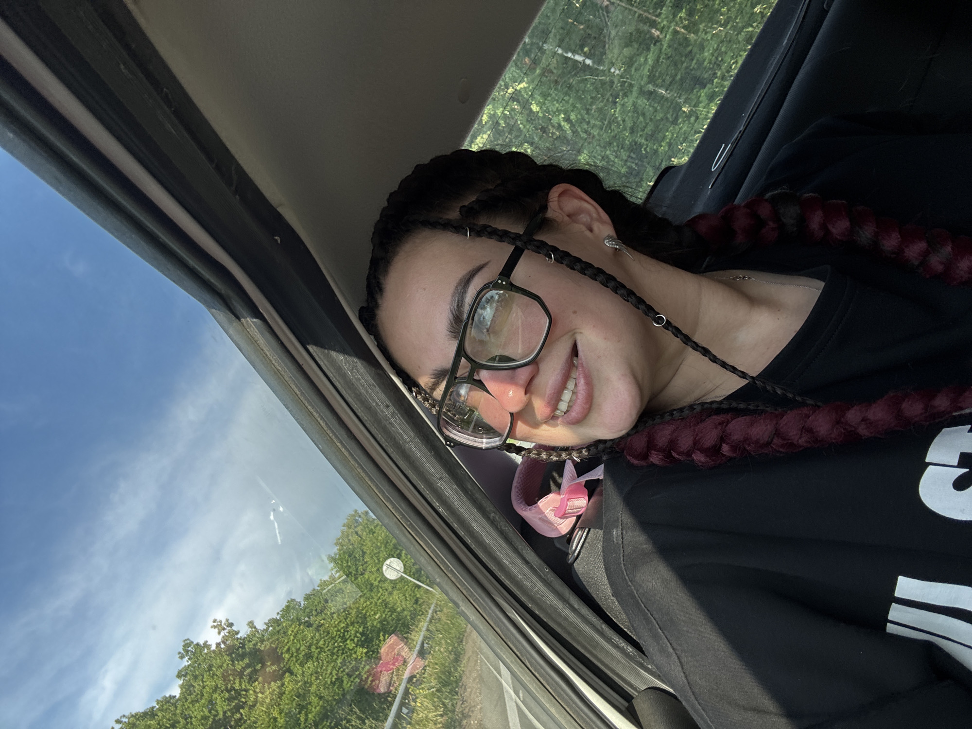 A young woman with dark braided hair and glasses is seen from the chest up, looking out of the window of a moving vehicle. She is wearing a black t-shirt. The background outside the window shows trees and a blue sky with white clouds. The interior of the vehicle, including the car door and window frame, is visible. The scene appears to be captured during daytime.